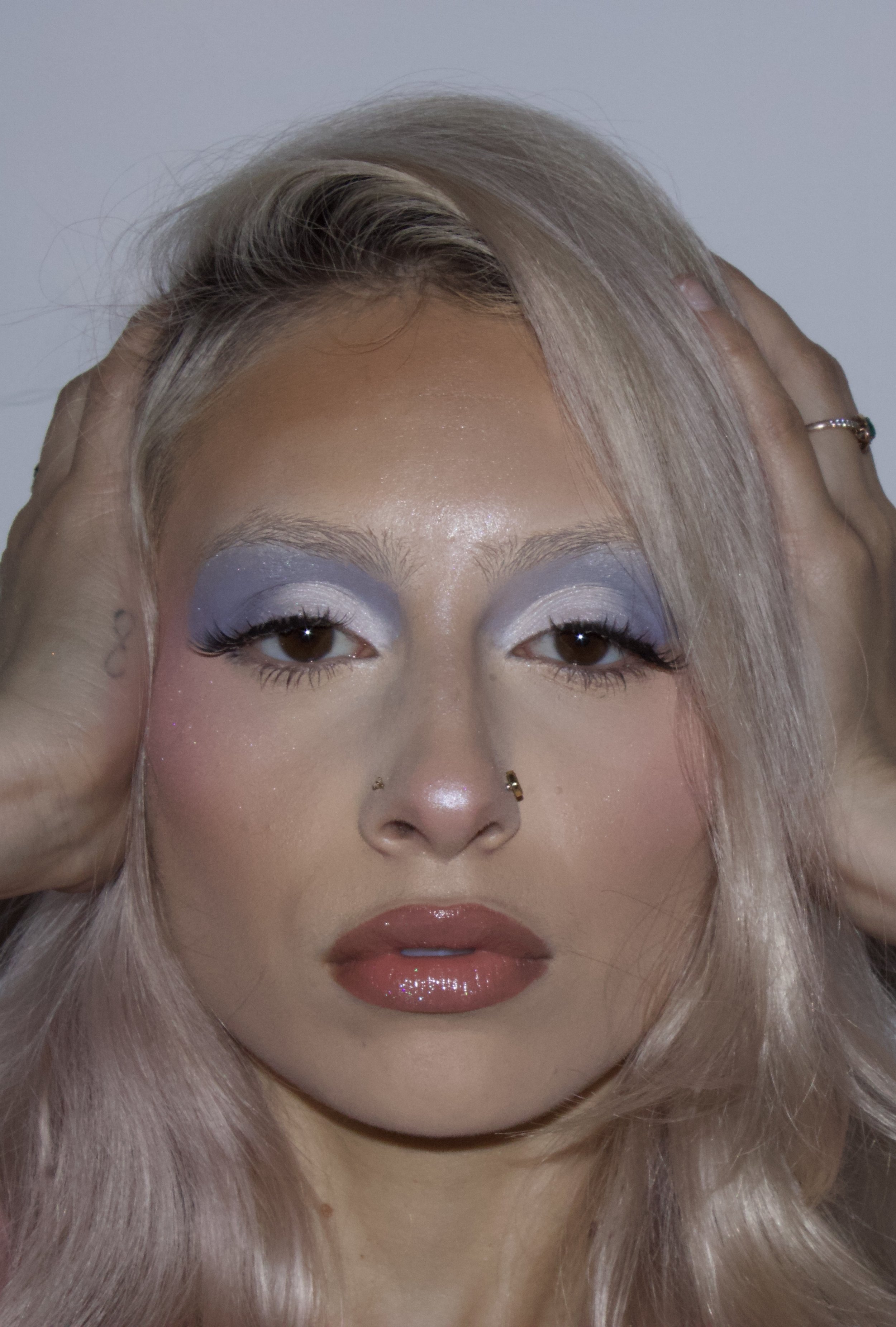 Close-up of a woman with platinum blonde hair, bold eyeshadow, and shiny lip gloss, holding her head with both hands.