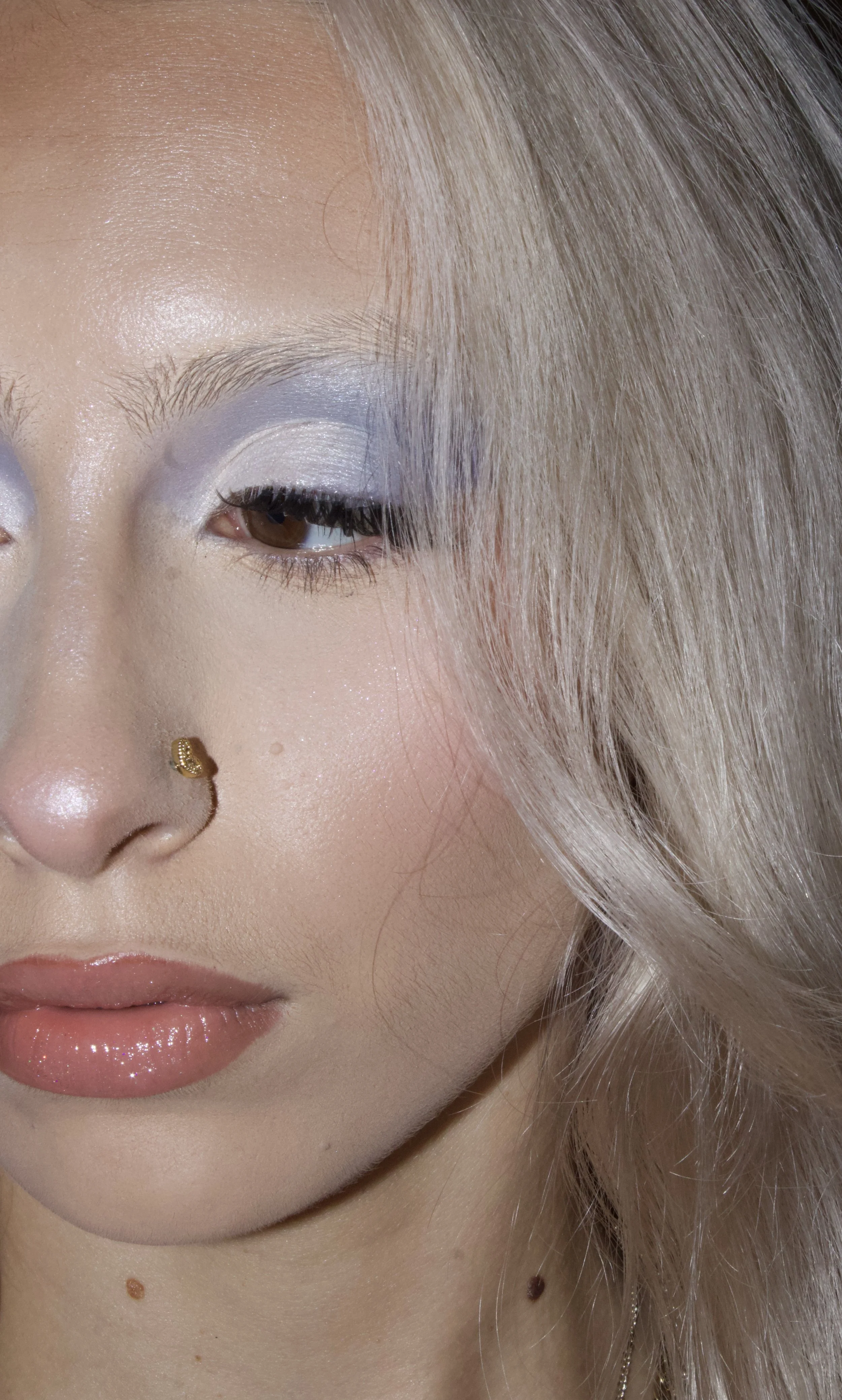 Close-up of a woman's face showing her eye makeup, lips, and a nose piercing, with platinum blonde hair and a few beauty moles on her neck.