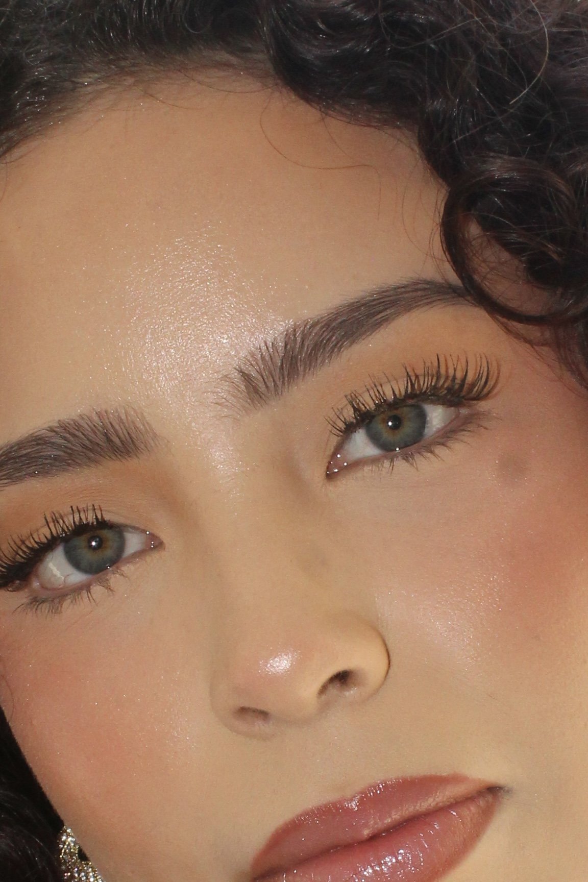 Close-up of a woman's face focusing on her eyes and forehead, with well-groomed eyebrows, long eyelashes, and makeup, including light eyeshadow and glossy lips.