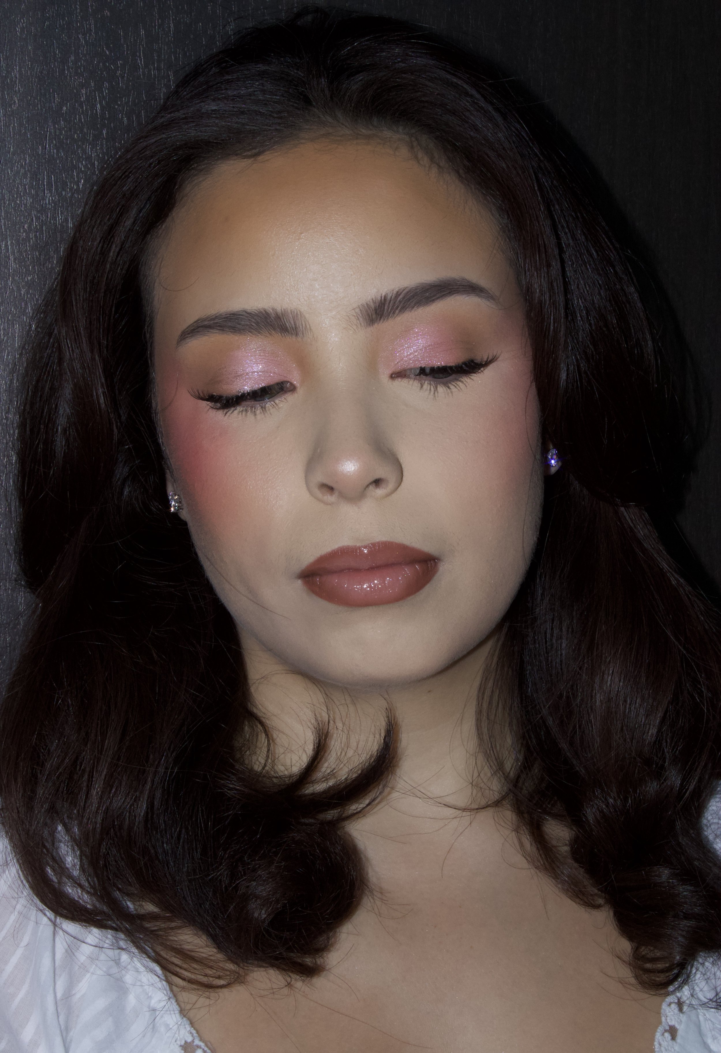 Close-up of a woman's face with makeup, dark wavy hair, and earrings, against a dark background.