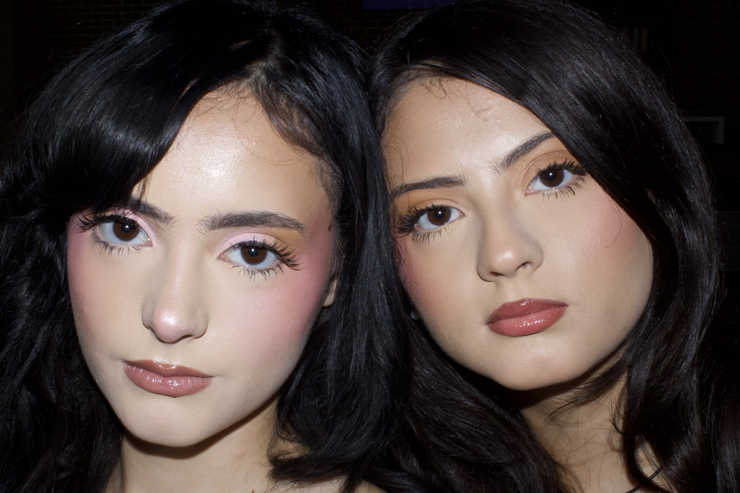 Close-up of two young women with black hair, brown eyes, and makeup, posing closely together indoors.