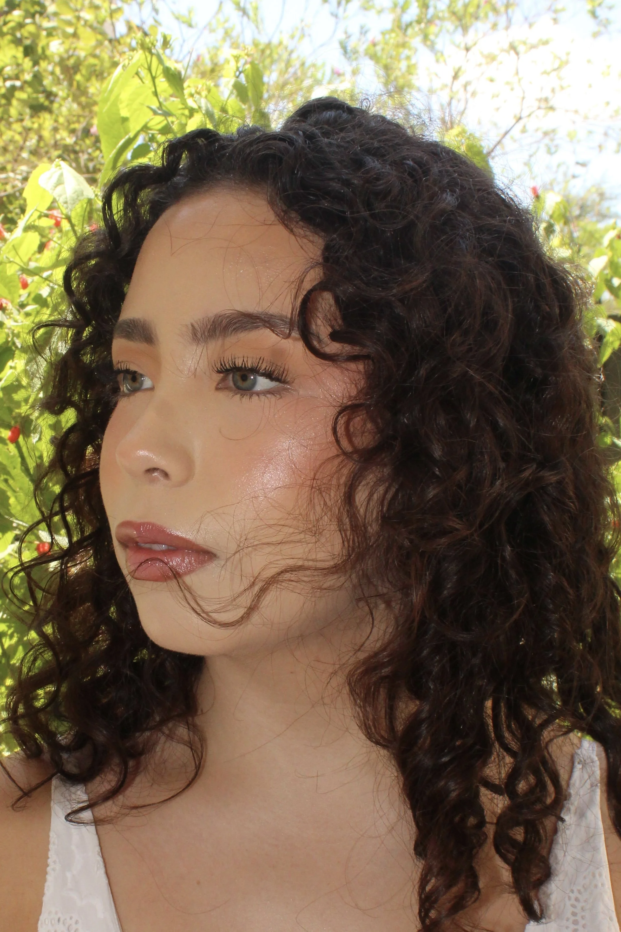 Close-up of a woman with curly dark hair, wearing a white top, outdoors with green foliage in the background.
