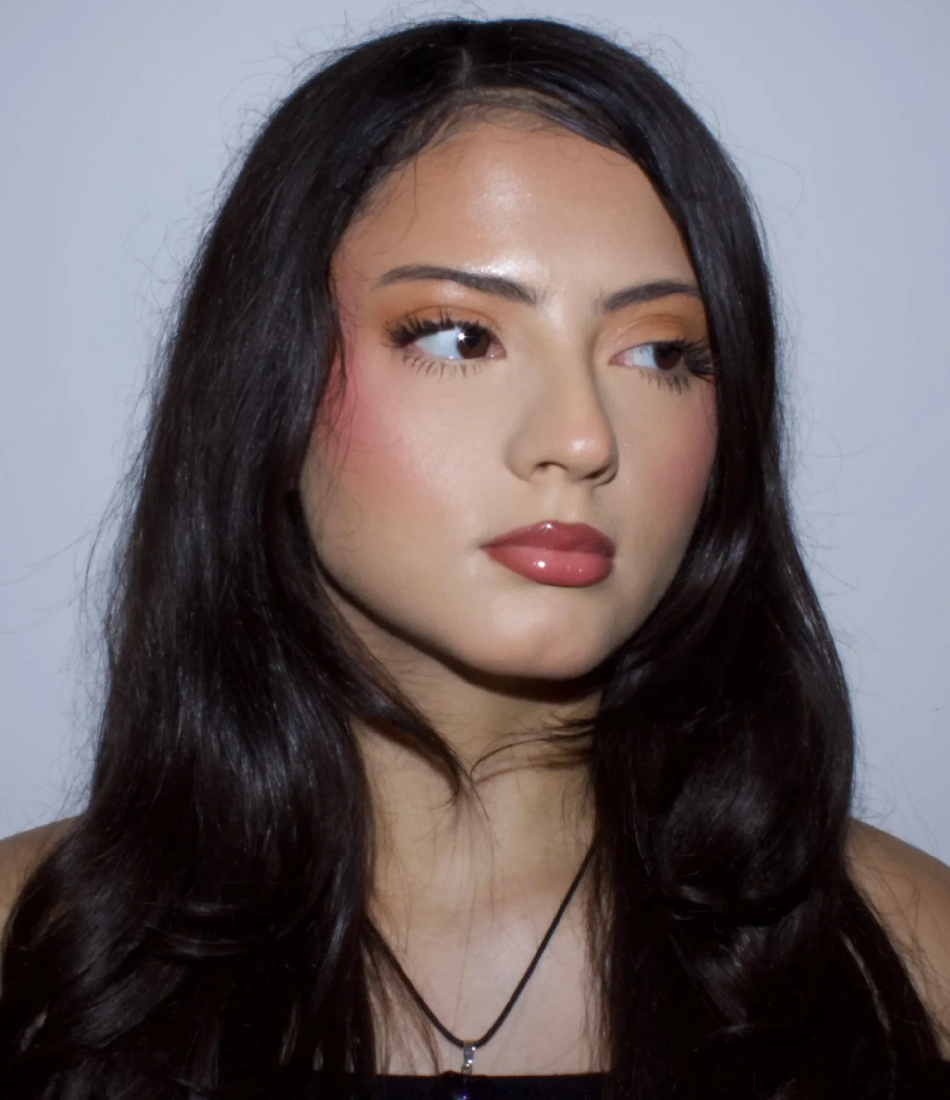 Close-up of a woman with long black hair, wearing makeup, a necklace, and earphones, against a plain background.