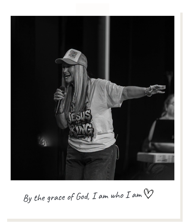 A woman wearing a baseball cap with the words "King of the Line" and a t-shirt that says "Jesus Is King," speaking into a microphone on stage. She has her right arm extended outward and is smiling, with a keyboard and keyboardist partially visible in the background.