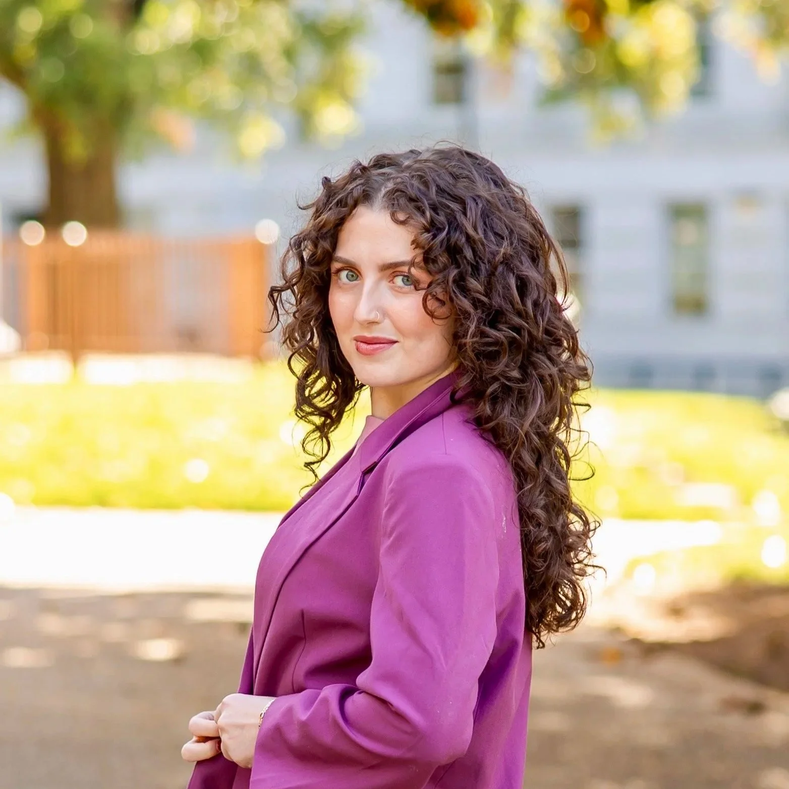 A woman with curly brown hair and blue eyes wearing a purple blazer standing outdoors in a park with trees and a building in the background.