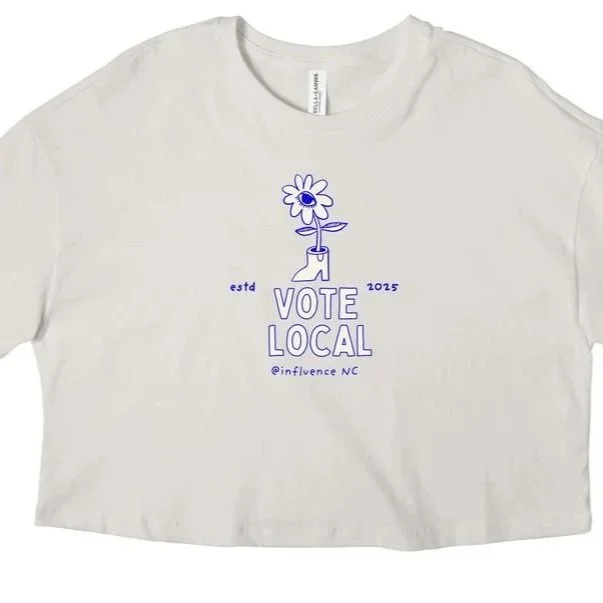 White t-shirt with purple graphic of a flower growing out of a boot, and text that reads "Vote Local," "estd 2025," and "@influence NC."