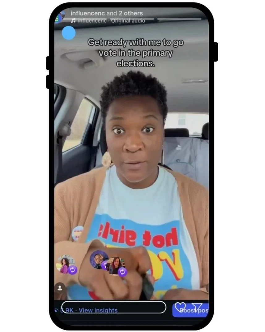 A woman with short curly hair and hoop earrings, wearing a colorful t-shirt and a tan cardigan, appears to be recording a video on her smartphone inside a vehicle. The screen shows her pointing towards the camera, with the message 'Get ready with me to go vote in the primary elections' displayed on top.