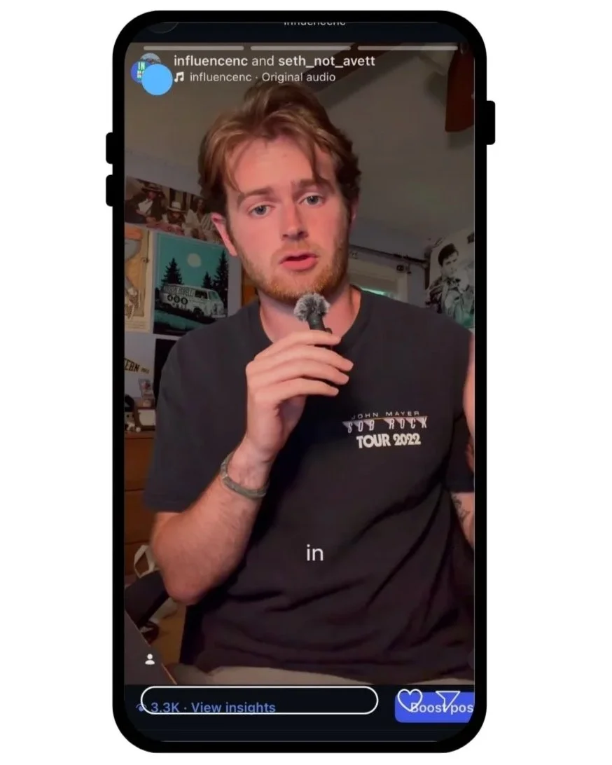 A man with red hair and a beard holding a microphone with a fluffy windscreen, sitting indoors in front of a background with posters and artwork, wearing a black t-shirt that says 'John Mayer Sub Roxy Tour 2022'.