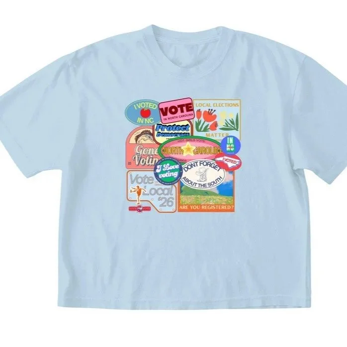 Light blue T-shirt with multiple colorful stickers and patches related to voting and elections.