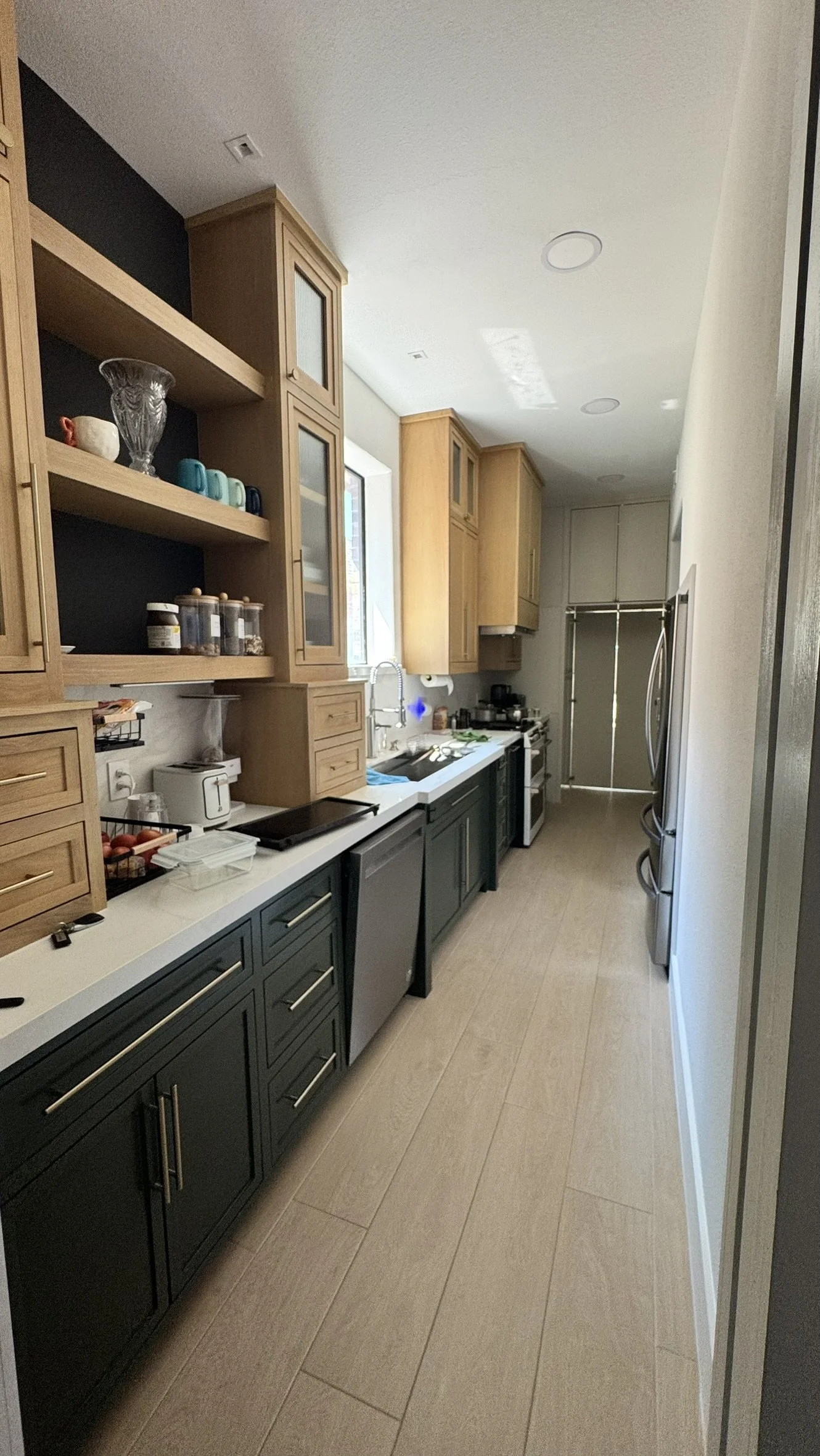 Modern kitchen with dark lower cabinets, wooden upper cabinets, and beige tile flooring. Appliances include a dishwasher, microwave, and refrigerator. Open shelving displays dishes and jars.