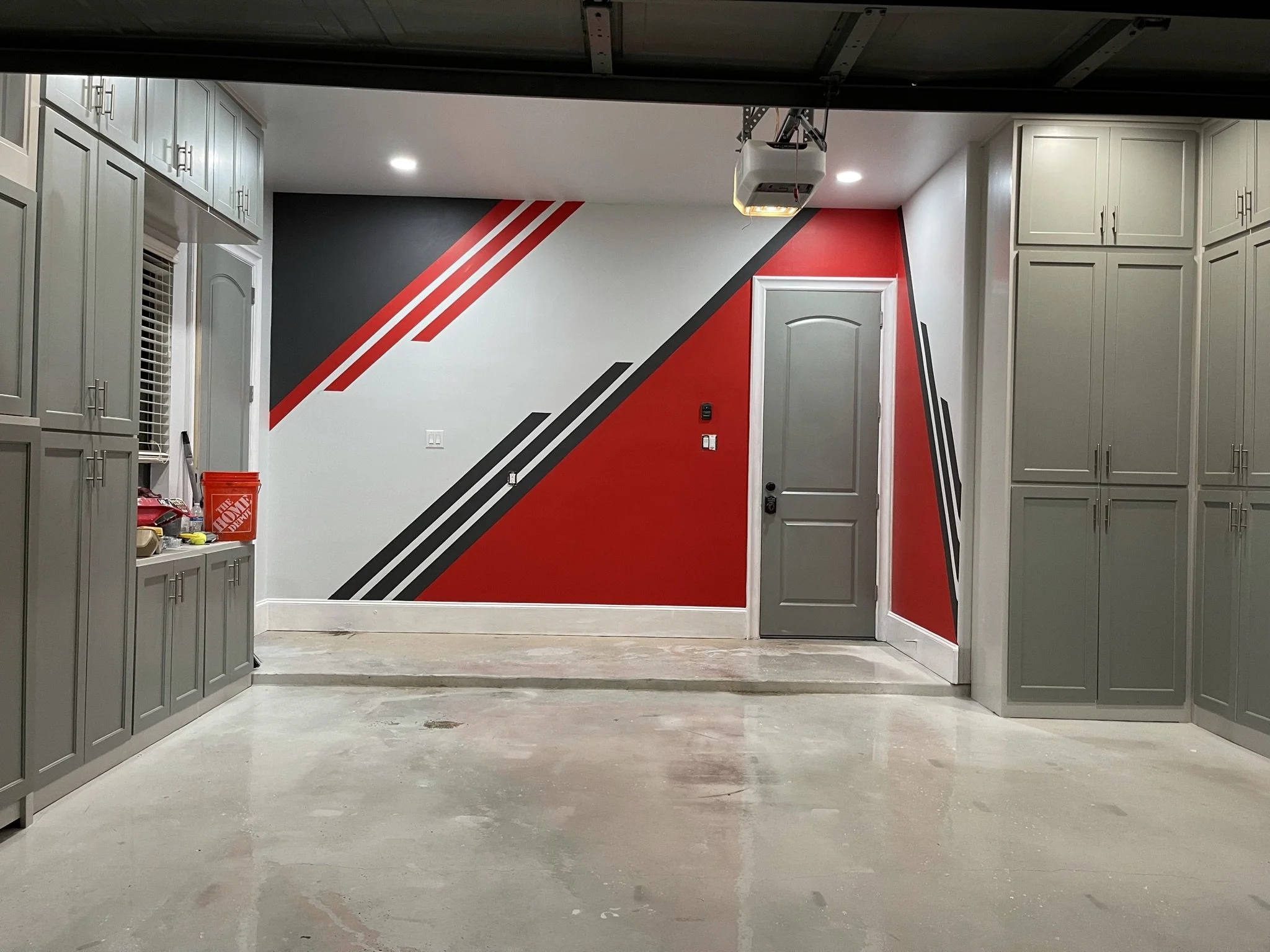 Garage interior with white cabinetry on both sides, a red and black geometric accent wall, and an automatic garage door opener installed on the ceiling.