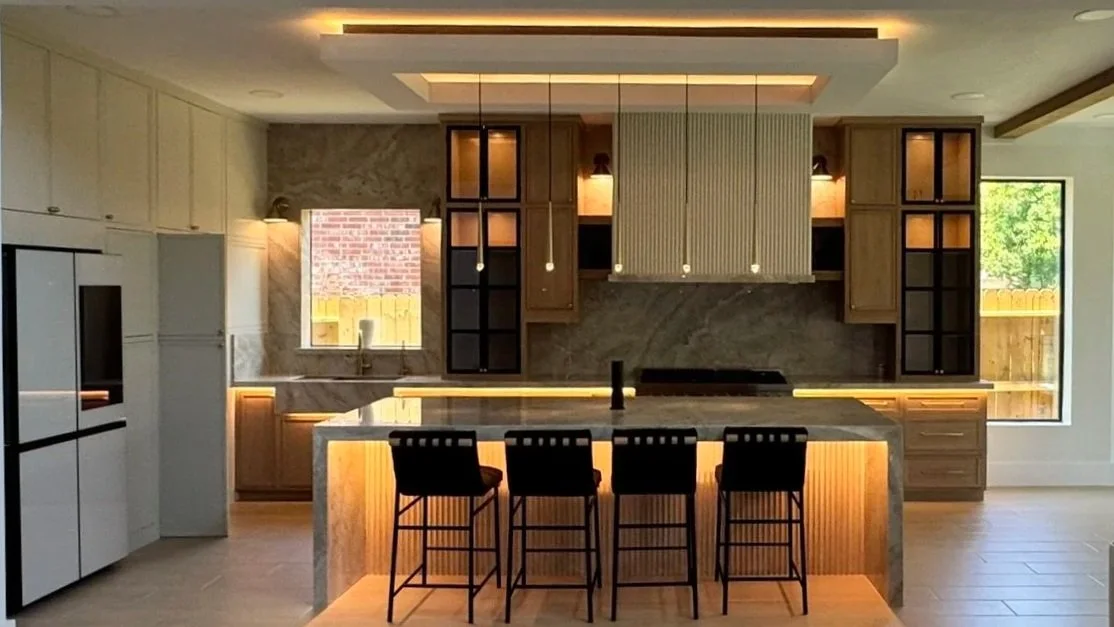 Modern kitchen with a marble island, four black bar stools, wooden cabinetry, and warm under-cabinet lighting. There are upper and lower cabinets, with a brick-patterned picture on the wall above the sink, and a window showing greenery outside.