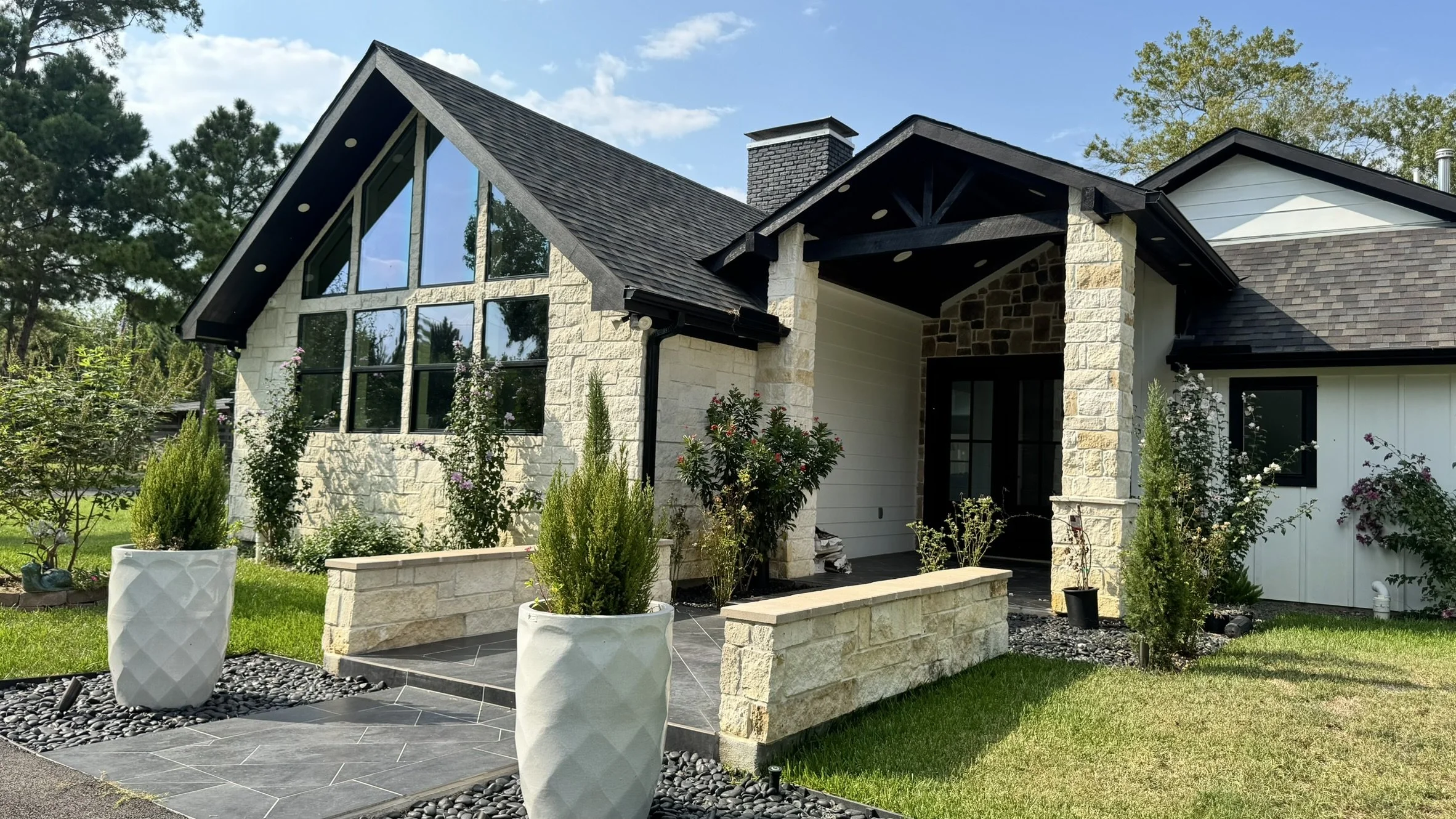 Modern house with large glass windows, stone and white siding exterior, black roof, and surrounding landscaping with potted plants and green lawn.