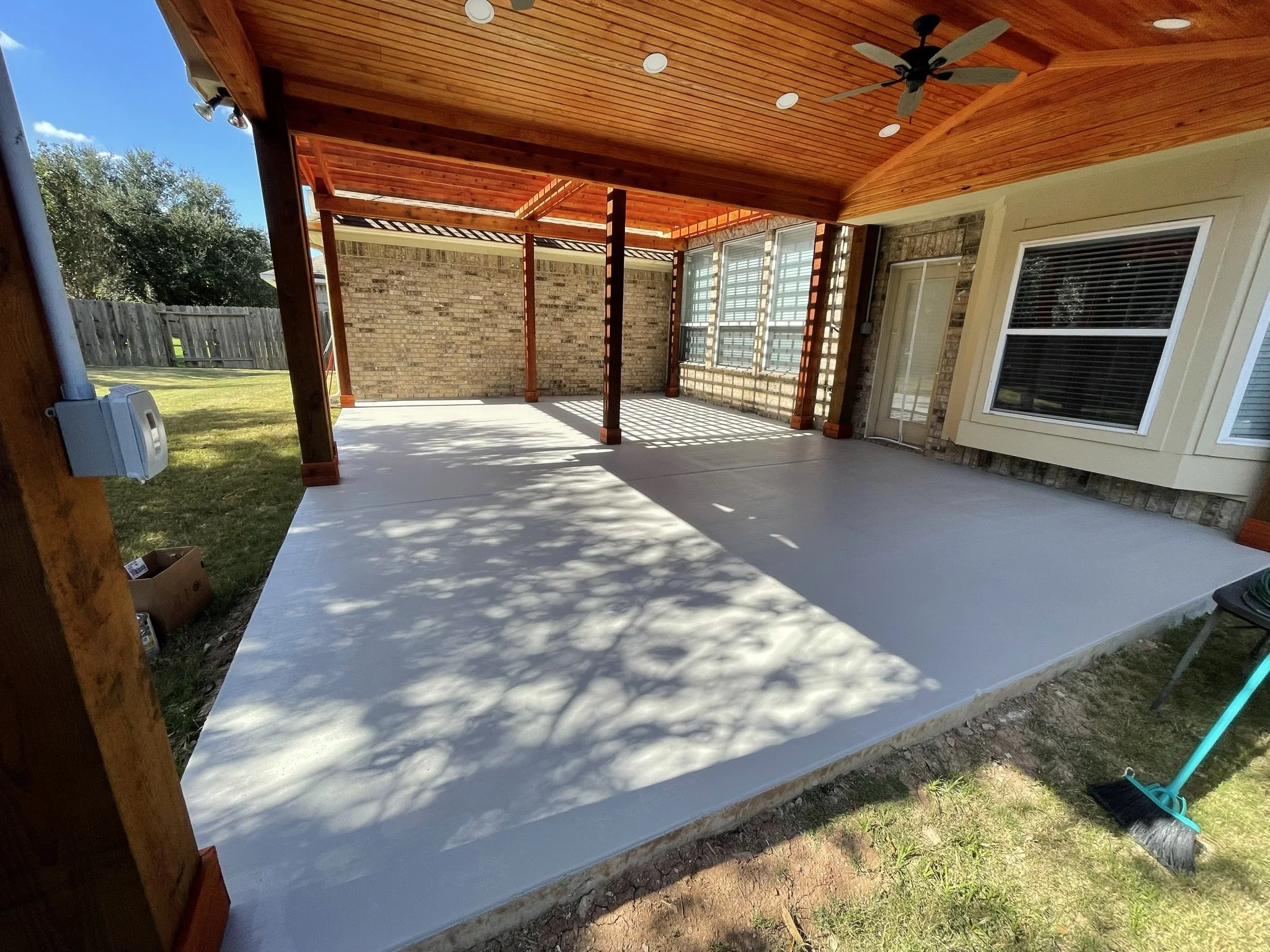 Newly poured concrete patio under a wooden pergola attached to a brick house. Surrounding grass yard and backyard fencing visible, with a blue sky overhead.