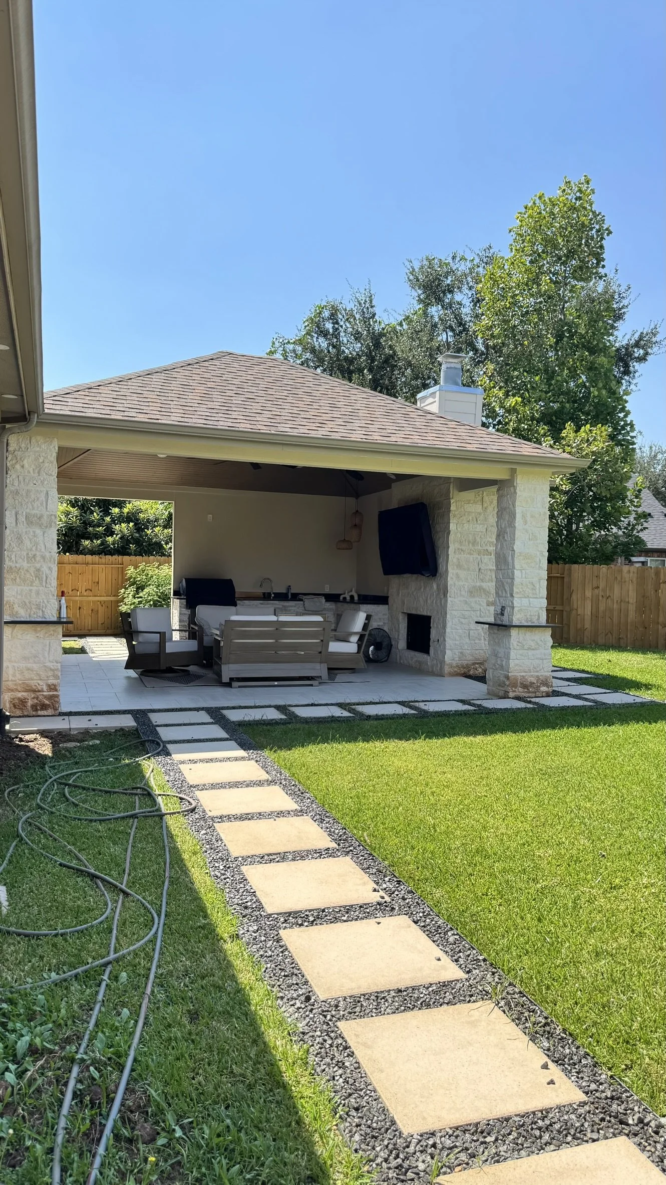 Backyard patio area with outdoor seating, a television mounted on stone wall, and a stone pathway leading to a fenced lawn.