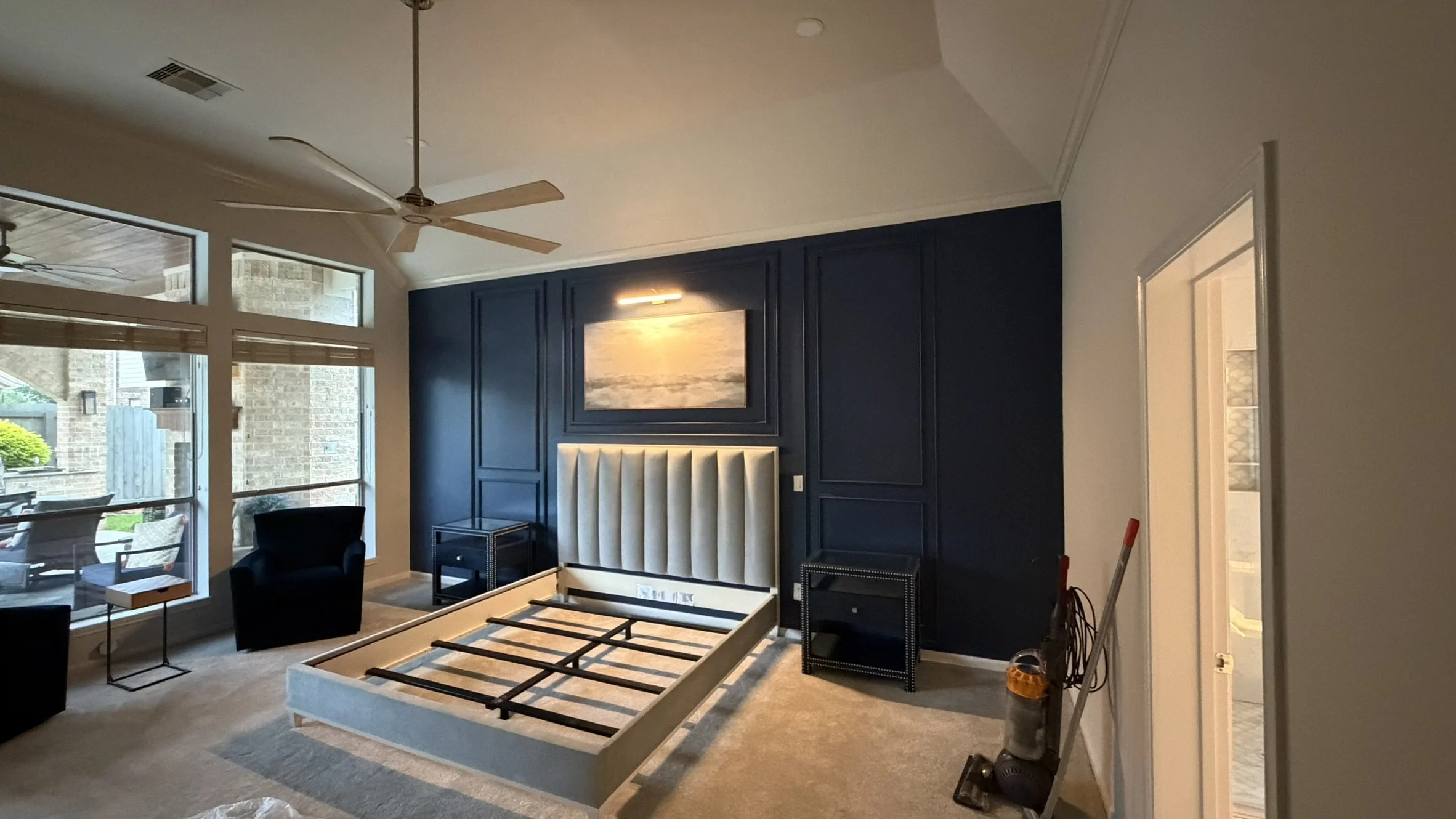 Living room with dark blue accent wall, large window, ceiling fan, and a beige headboard against the wall. The room appears to be in the process of furniture assembly.