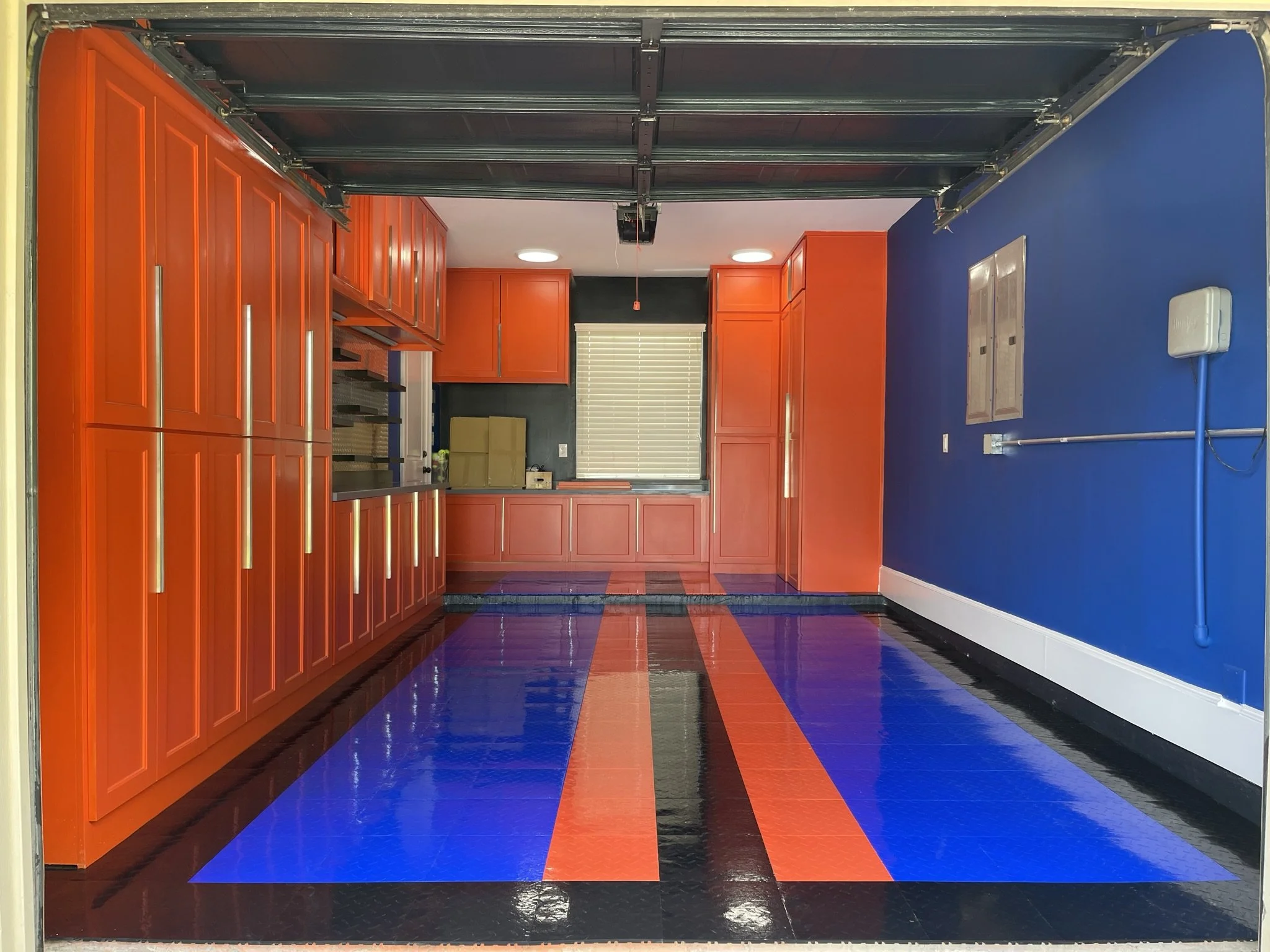 Open garage door revealing a room with orange cabinets, blue and black floor, and a blue and white wall. There is a window with blinds, ceiling lights, and a cardboard box on the counter.