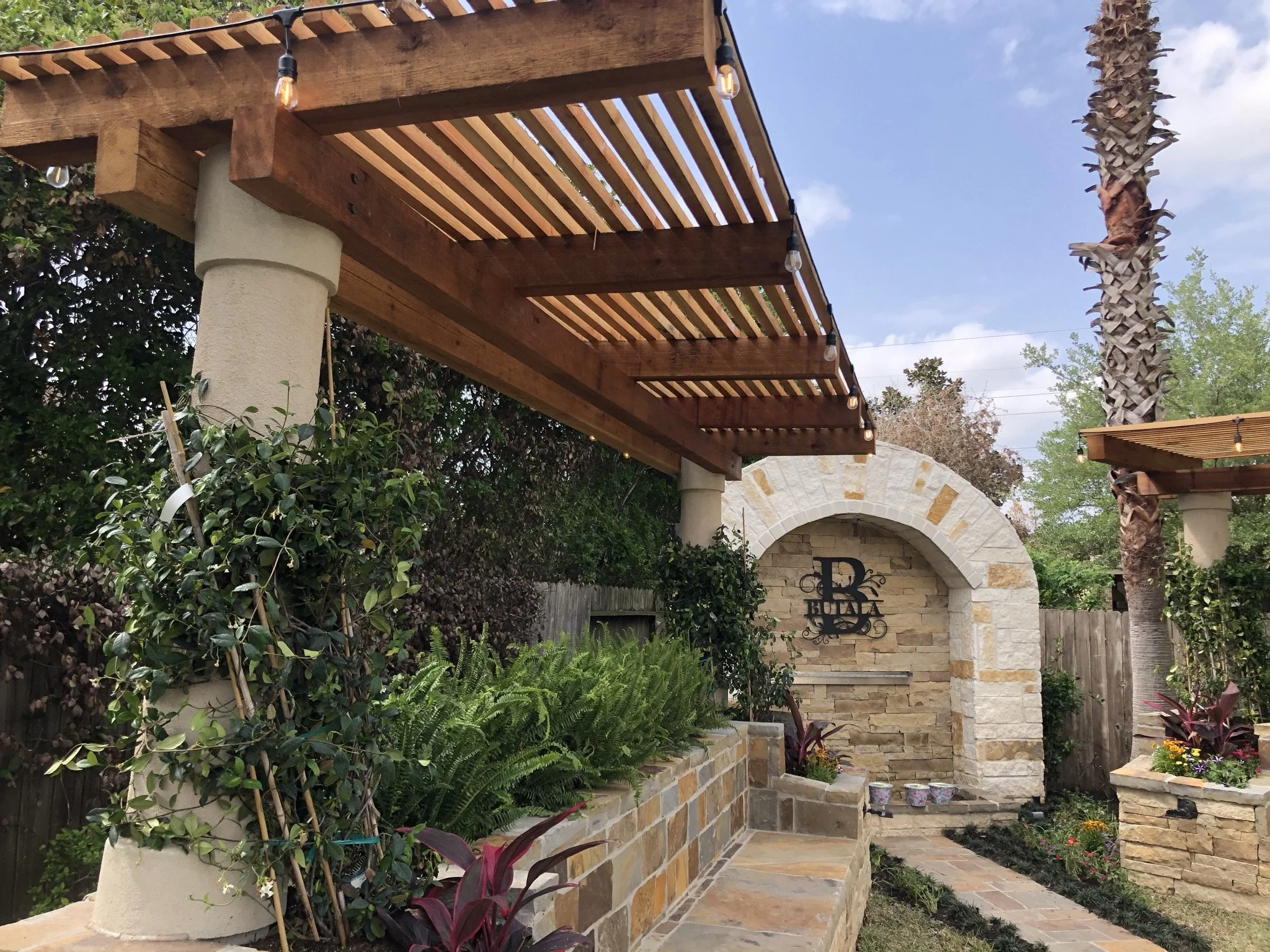 A landscaped backyard with a stone archway, a wooden pergola with string lights, lush greenery, and potted plants.