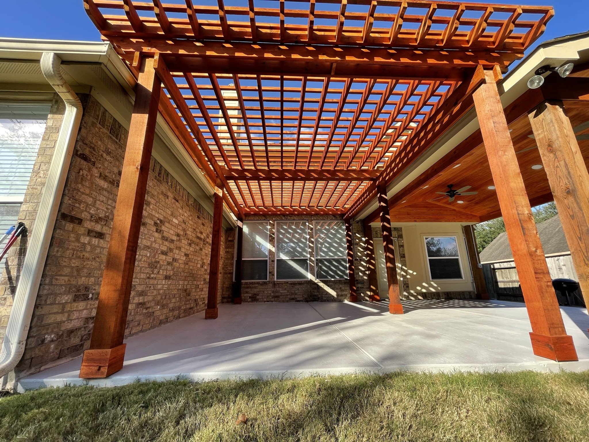 View of a backyard patio area with a wooden pergola structure, brick walls, windows, and outdoor ceiling fans, with clear blue sky in the background.