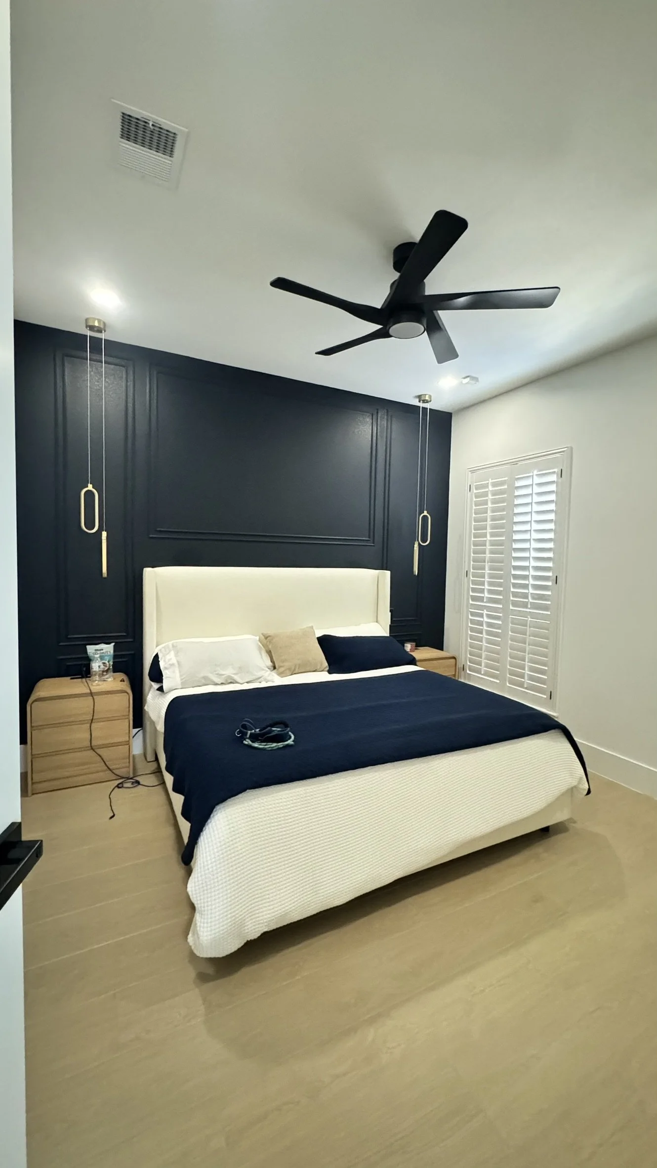 Modern bedroom with a beige upholstered bed, navy blue bedding, black pillows, and a beige pillow, two wooden nightstands, hanging pendant lights, a ceiling fan, a window with white shutters, and a vent on the ceiling.