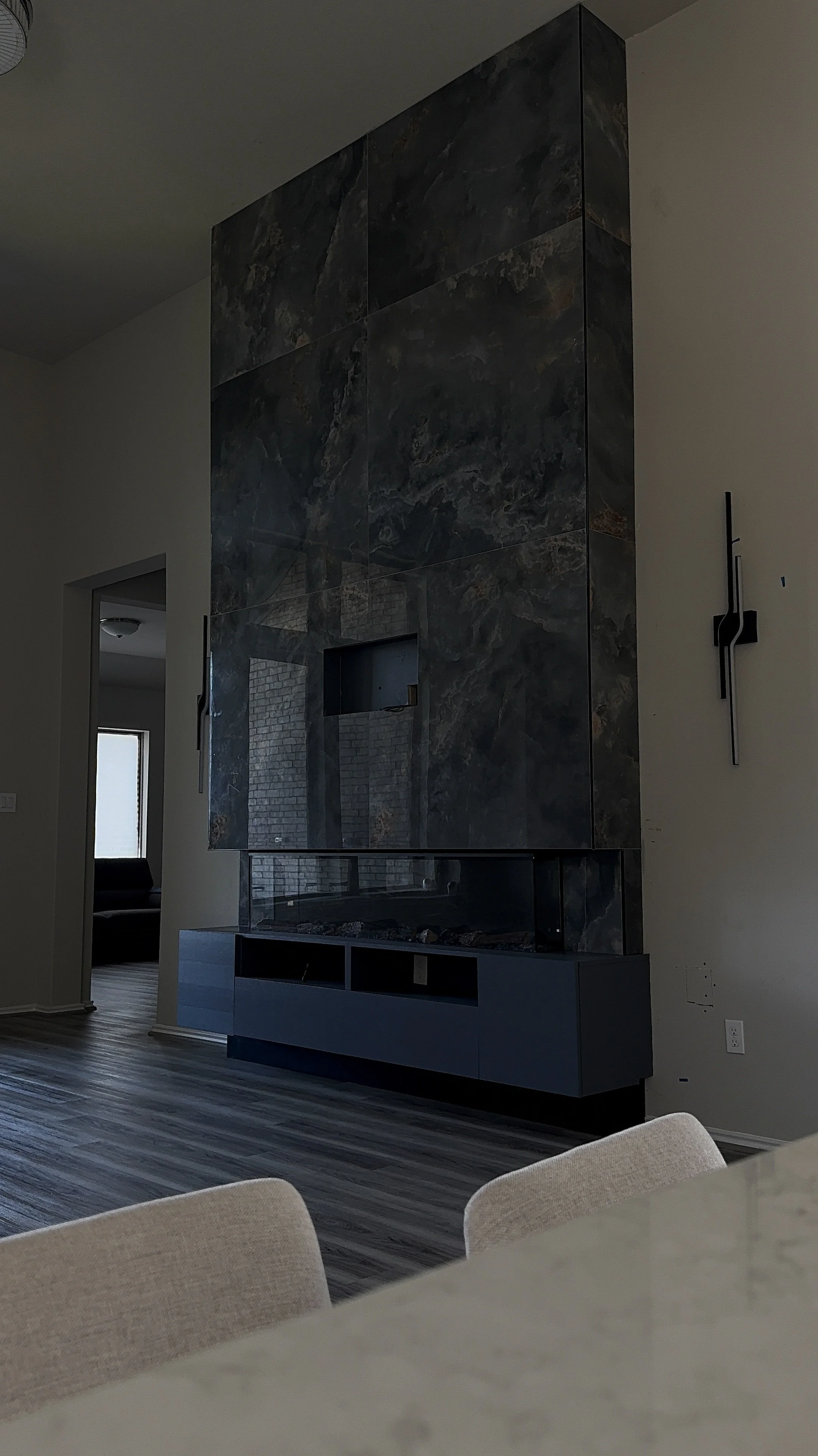 Interior of a modern living room with a dark stone fireplace, hardwood floors, and a doorway leading to another room. Part of a chair and a table are visible in the foreground.