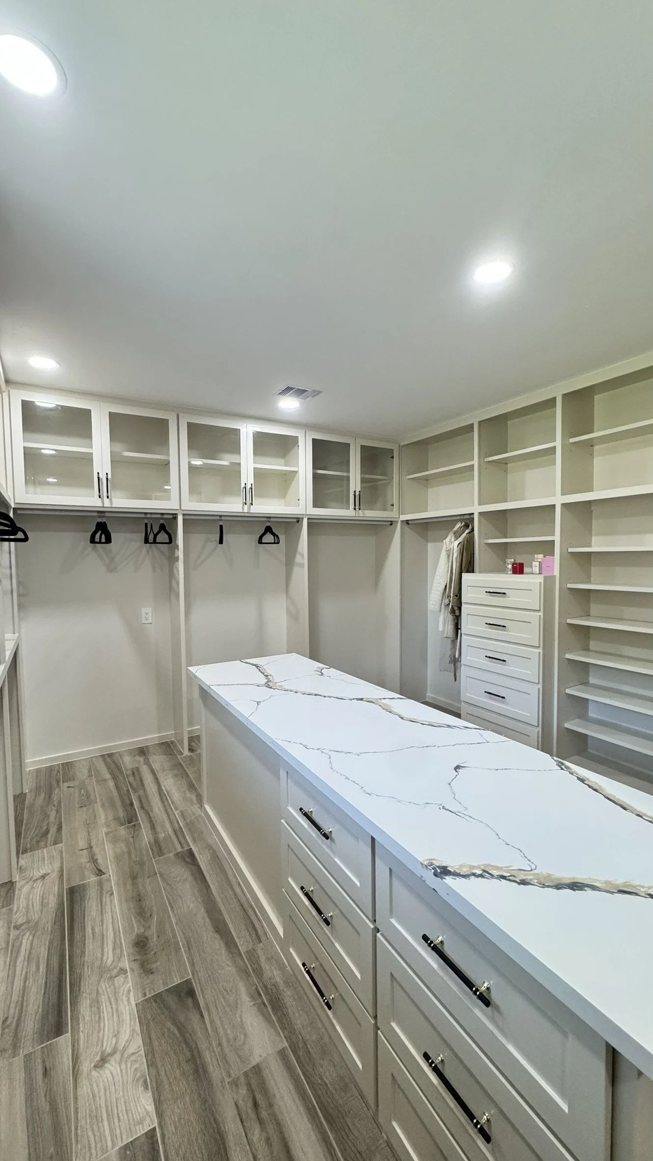 Empty walk-in closet with white cabinets, glass-fronted upper cabinets, black handles, open shelves, hanging rods, built-in drawers, marble countertop island, and wood-look flooring.