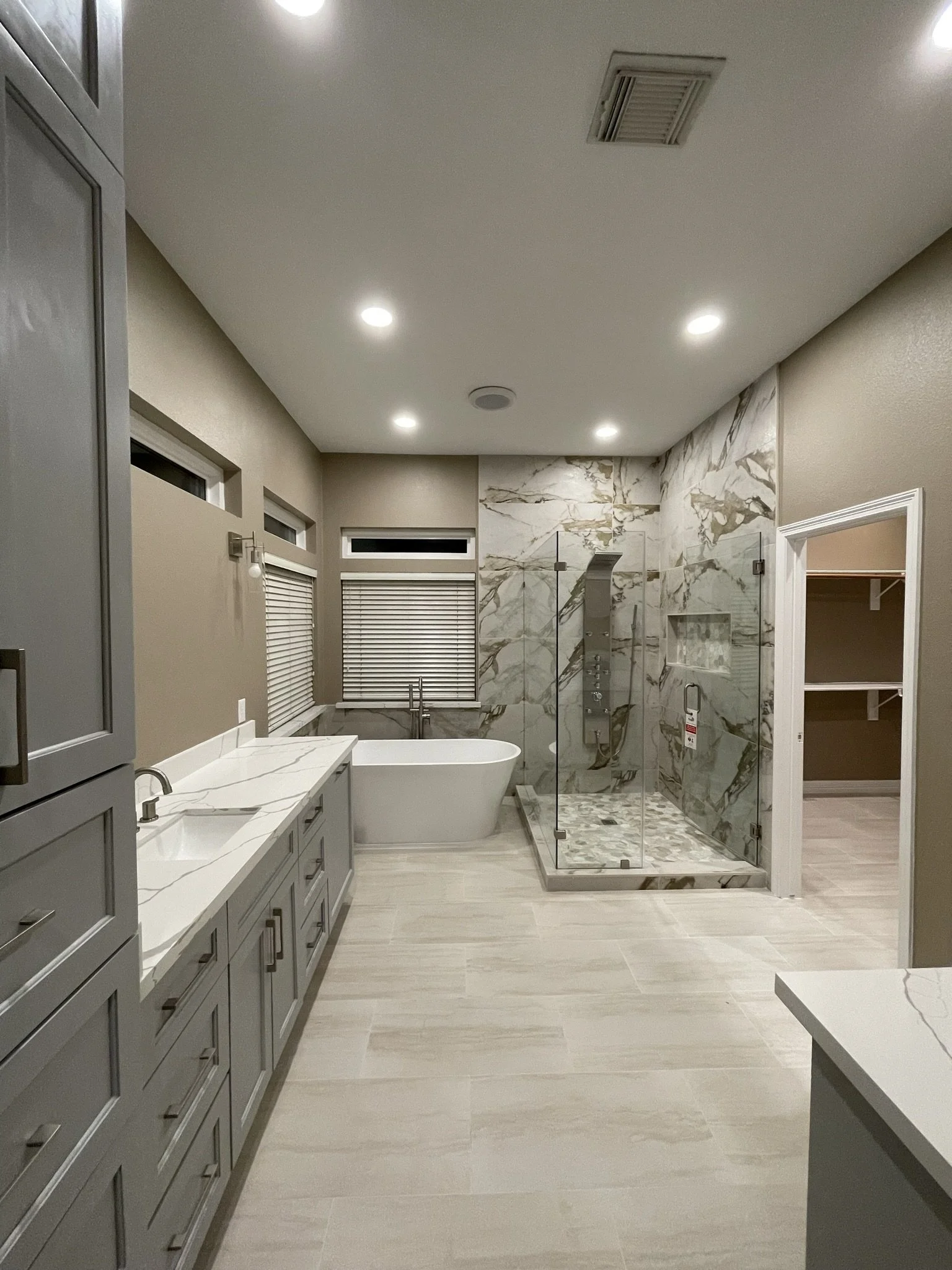 Modern bathroom with gray cabinets, white countertops, marble accent wall, walk-in shower with glass door, freestanding bathtub, and beige wood-look floor tiles.
