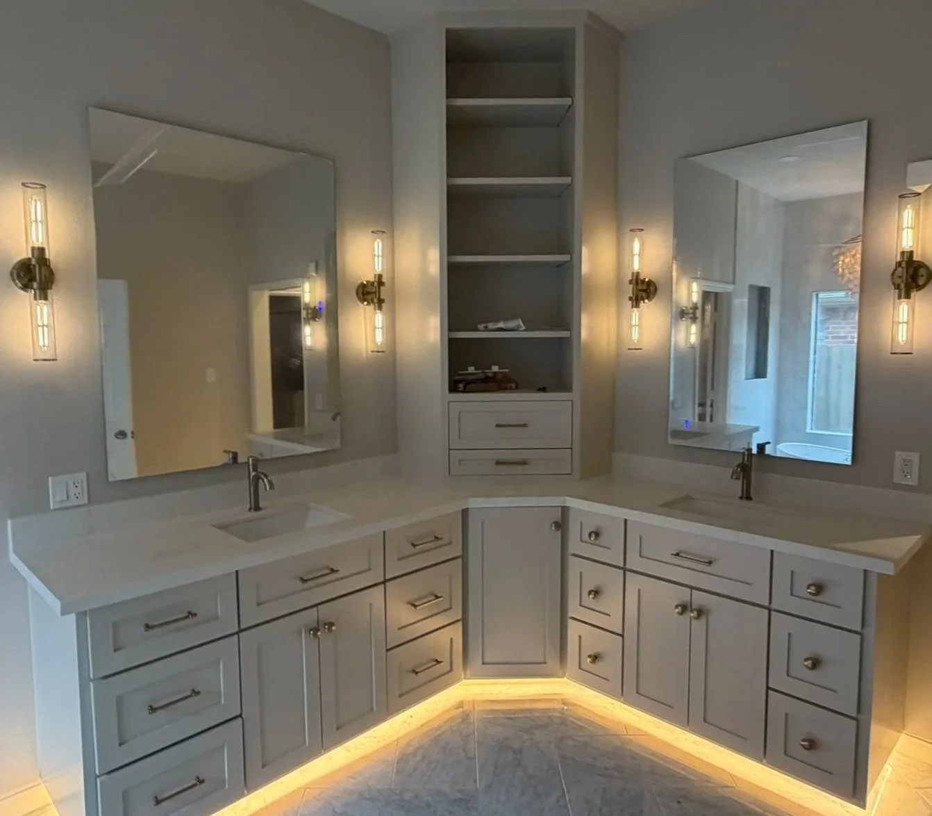 Modern bathroom vanity with dual sinks, white cabinets, large mirrors, wall-mounted light fixtures, and a central open shelving unit