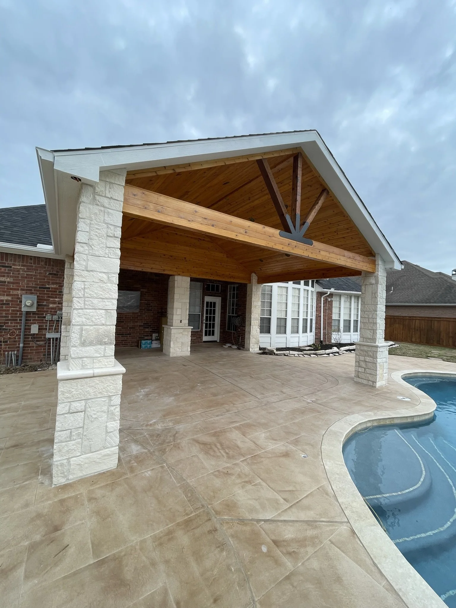 Backyard with a covered patio area, a swimming pool, and a stone paved deck, with a cloudy sky overhead.