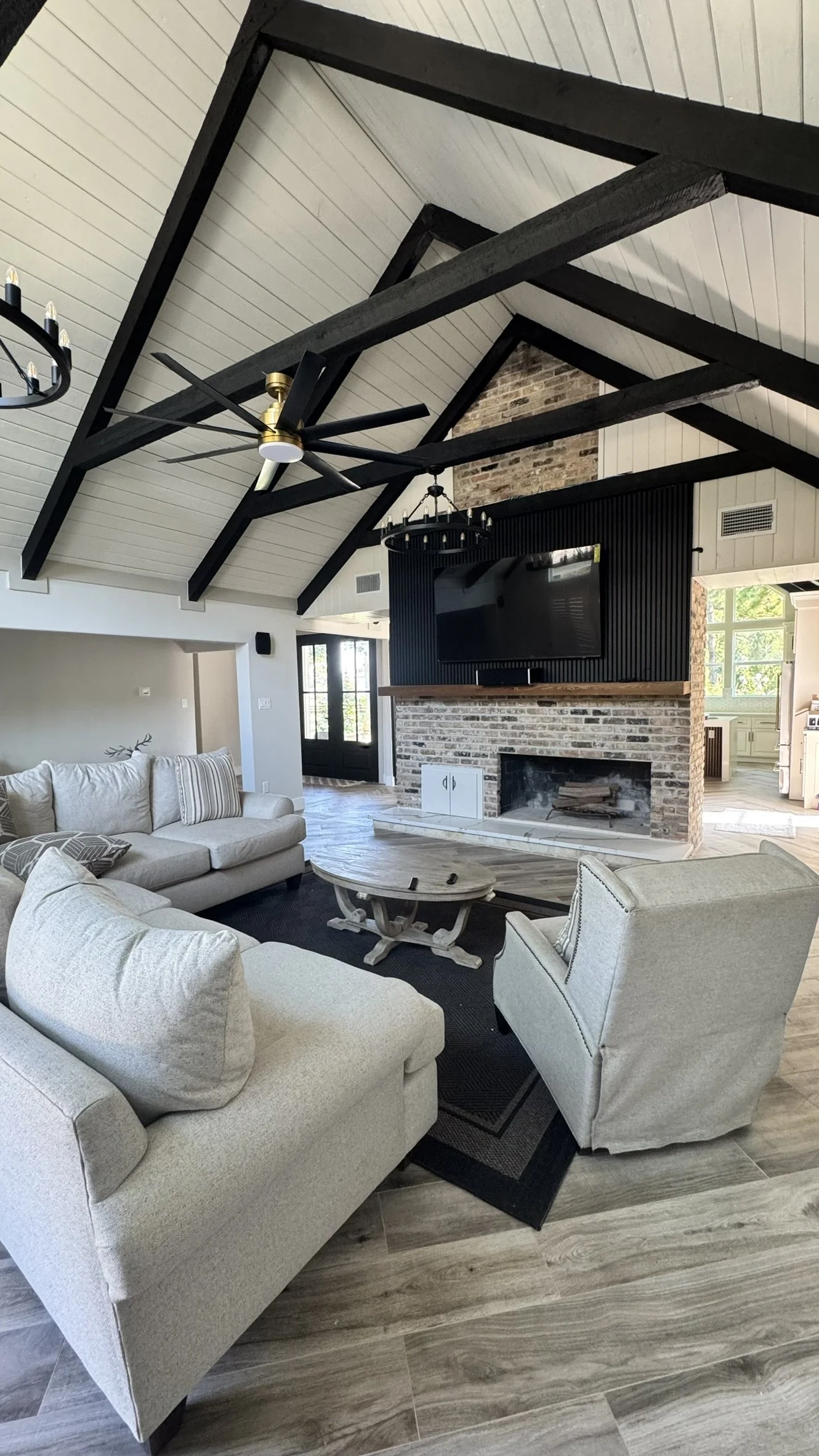Open living room with white sofas, a wooden coffee table, and a brick fireplace with a mounted TV. Black ceiling beams, a ceiling fan, and chandeliers are visible. A door and windows allow natural light.