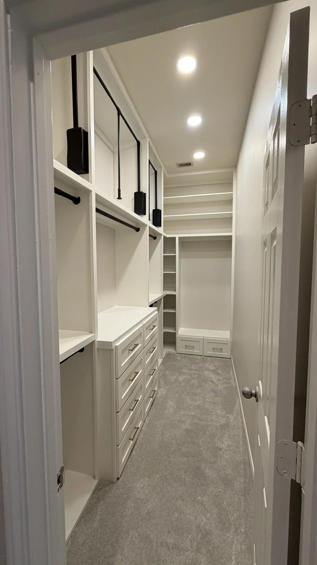 Empty walk-in closet with white built-in shelves, drawers, and hanging rods, gray carpet, and recessed ceiling lights.
