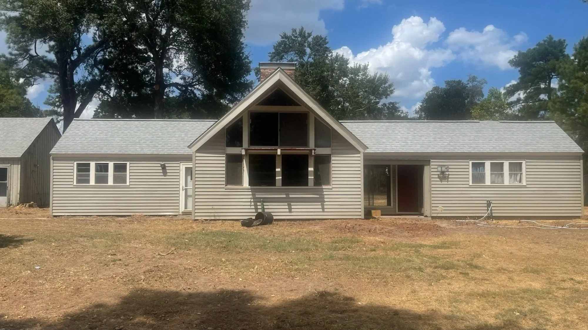 A house under construction with beige siding and a peaked roof, surrounded by trees, with some construction materials and tools visible in the yard.
