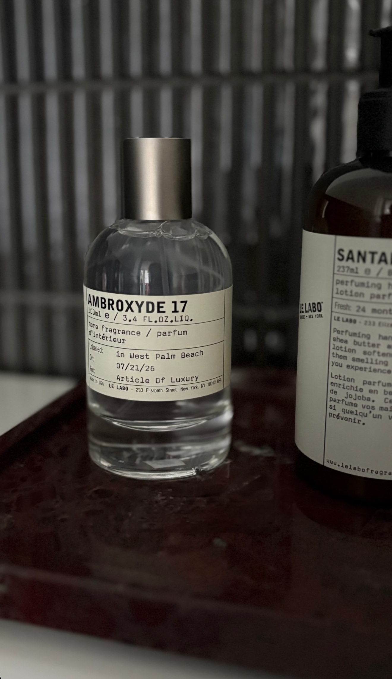A clear bottle labeled 'Ambroxide 17' with a silver cap, placed on a dark surface. Behind it, a partially visible brown bottle with a white label, and a metal background.