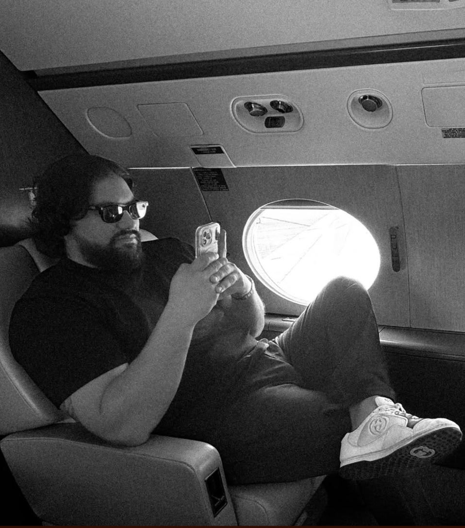 A man with dark hair, beard, and glasses lying in an airplane seat, wearing a black shirt, dark pants, white sneakers, and sunglasses, looking at his phone with a window behind him showing the sky outside.