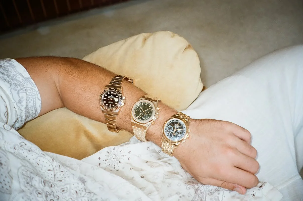 A person wearing three gold wristwatches on their left arm, resting on their lap while sitting on a beige cushion. The person is dressed in a white, patterned long-sleeve shirt and white pants.