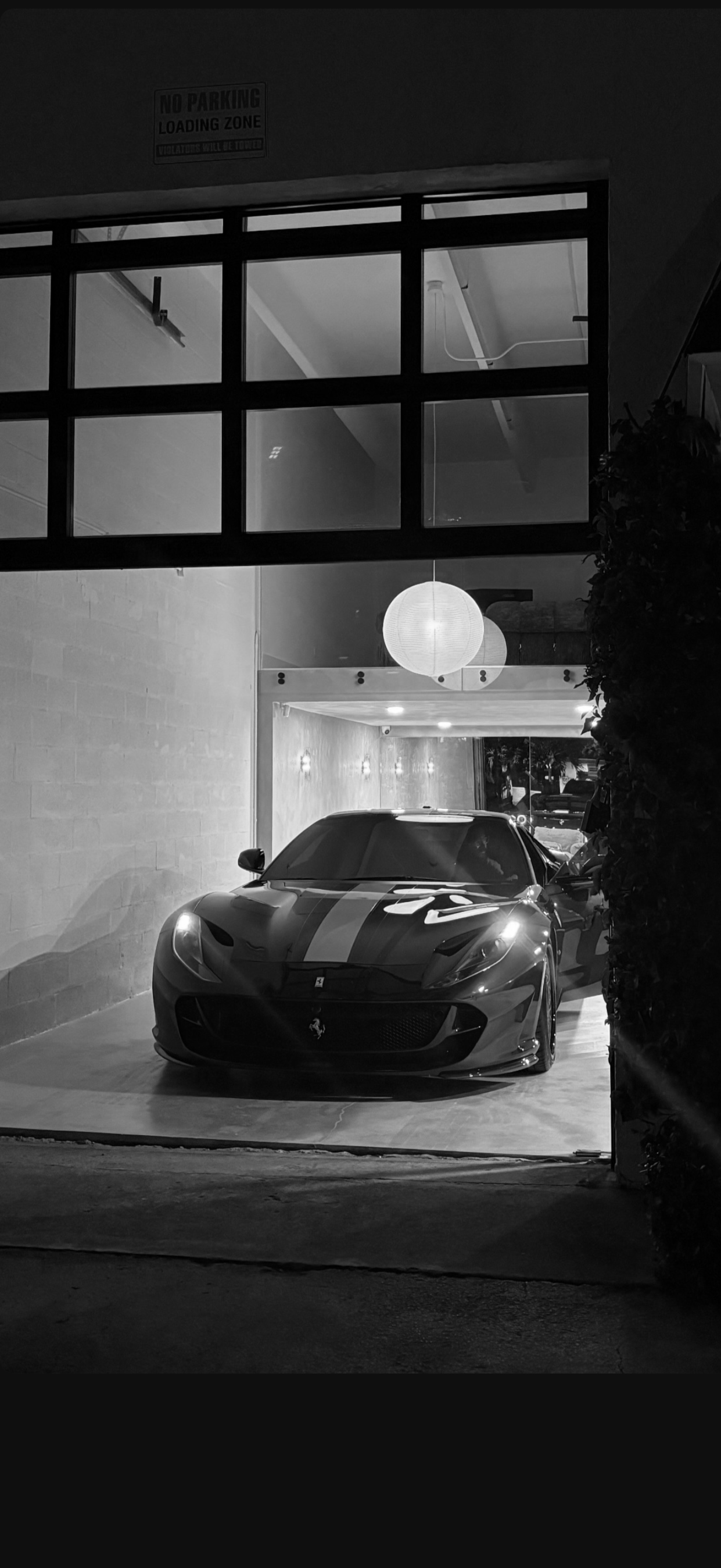 A black sports car parked inside a modern, well-lit garage at night, with a large window showing the interior and exterior lighting, and a sign reading 'No Parking Loading Zone' above.