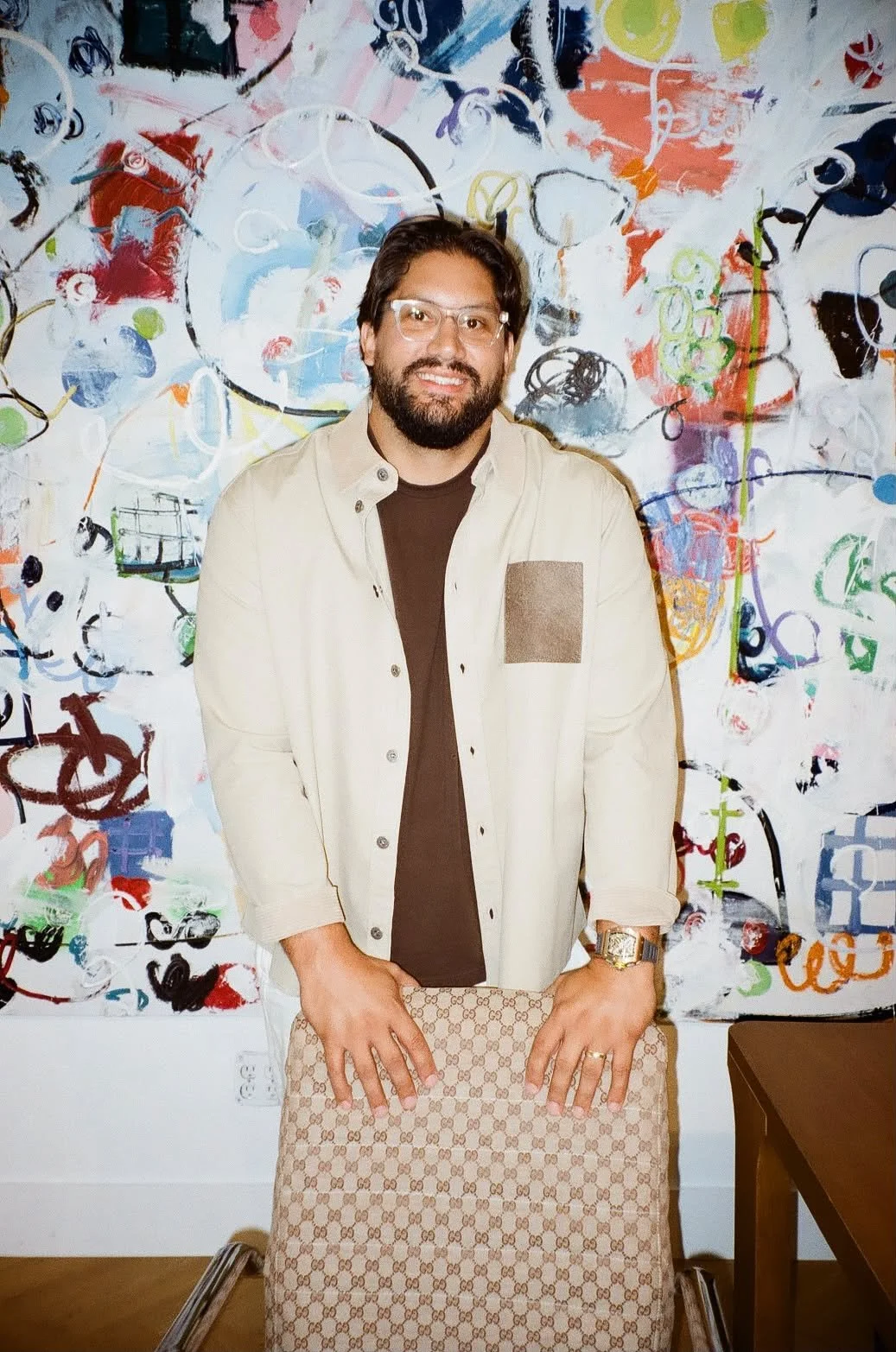 Man with glasses, dark hair, and beard standing in front of an abstract colorful painting, wearing a beige jacket, brown shirt, and holding a patterned handbag.