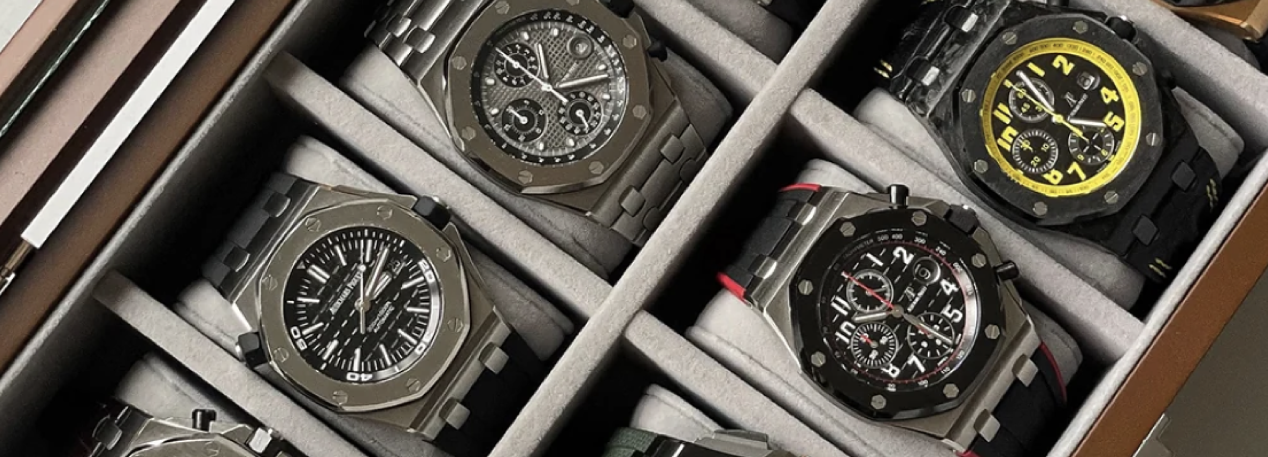 Six luxury wristwatches in a watch box, three with black faces and three with colored accents.