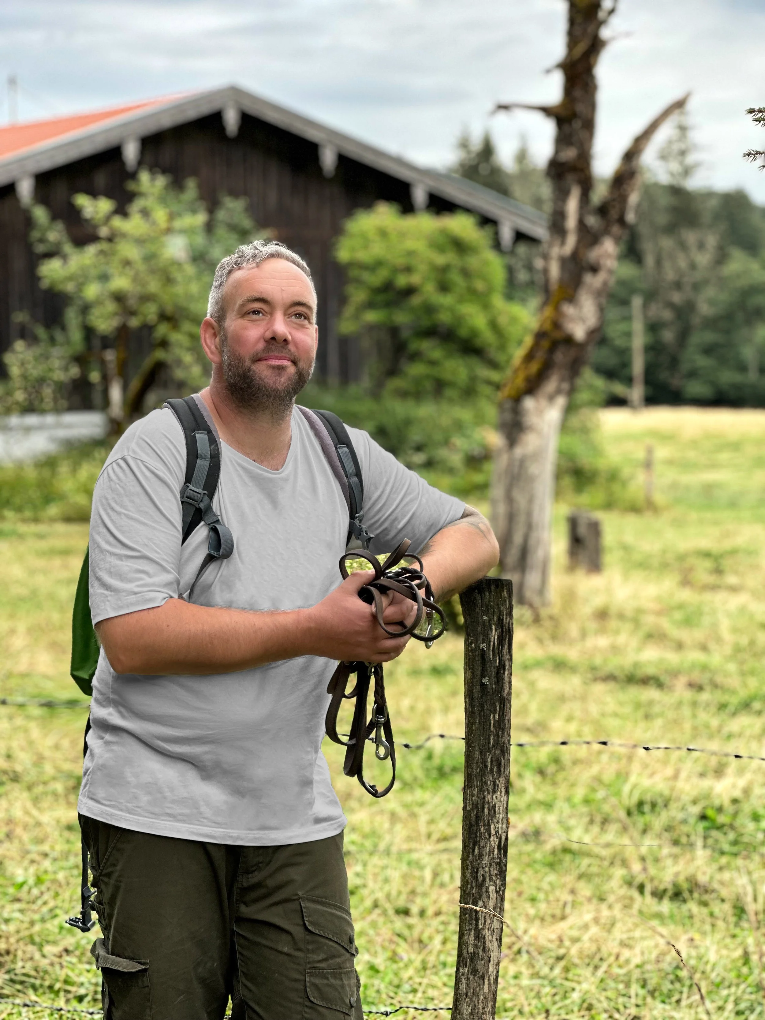 Ein Mann mit grauem Haar und Bart steht im Freien, trägt ein graues T-Shirt, einen Rucksack und hält Hundeleinen in den Händen, im Hintergrund Wald und eine Holzhütte.