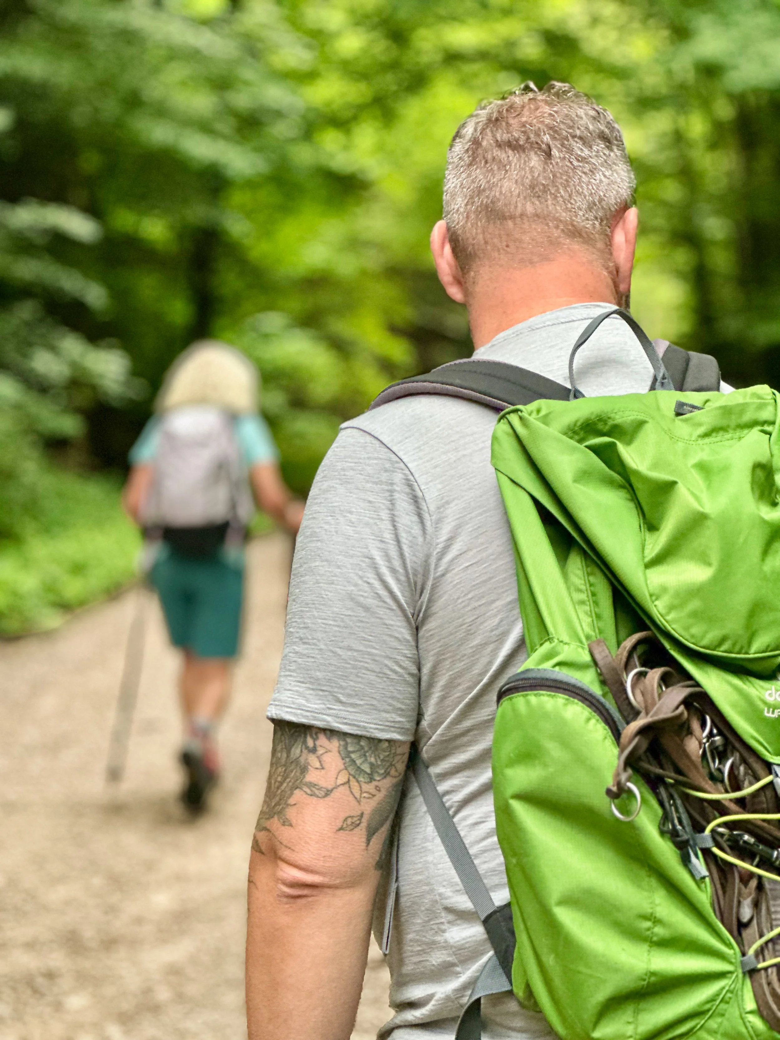 Ein Mann mit kurzen, grauen Haaren und tätowiertem Arm trägt einen grauen T-Shirt und einen grünen Rucksack, während er auf einem Waldweg geht. Im Hintergrund ist eine Frau mit einem hellen Rucksack, die weiter vorne läuft, zu sehen.