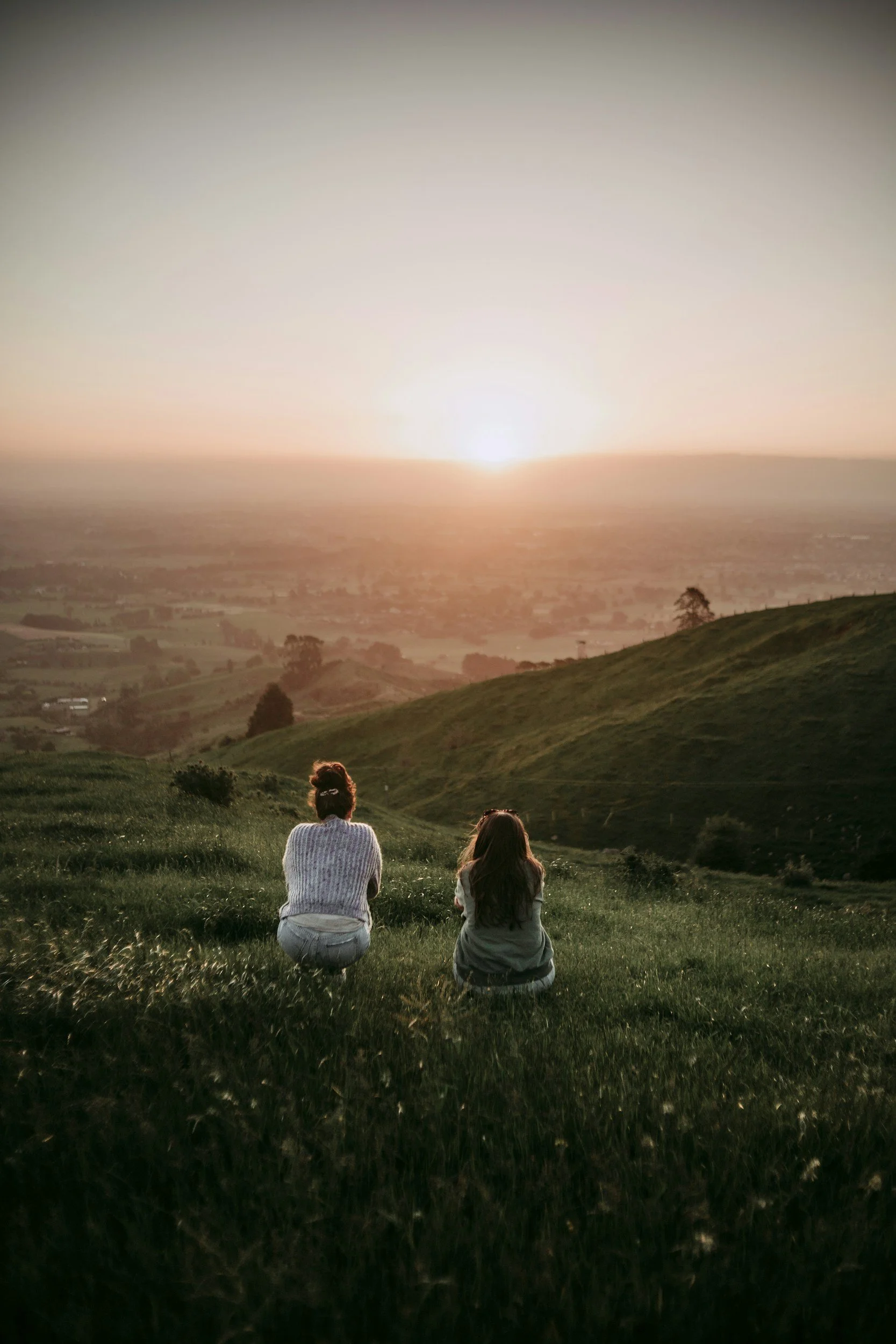 Two girls sitting on a grassy hill watching a sunset over the countryside.