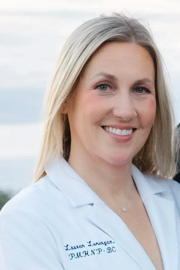 A woman with blonde hair and blue eyes smiling, wearing a white lab coat with a name embroidered on it, outdoors with a cloudy sky in the background.