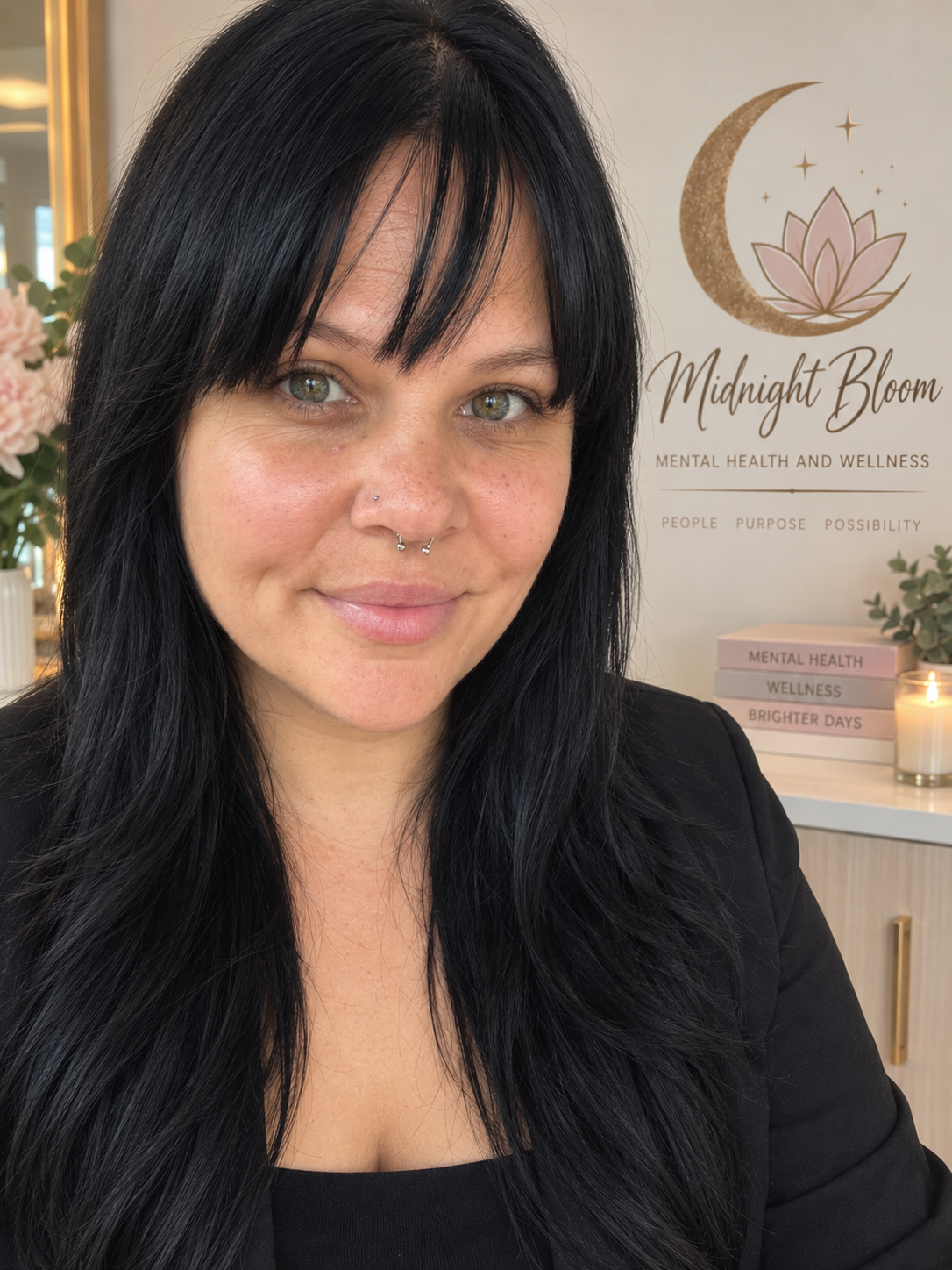A woman with black hair and a septum piercing smiling inside a wellness center. Behind her, a sign reads 'Midnight Bloom Mental Health and Wellness' with a lotus flower and moon logo, and books titled 'Mental Health,' 'Wellness,' and 'Brighter Days' on a table.