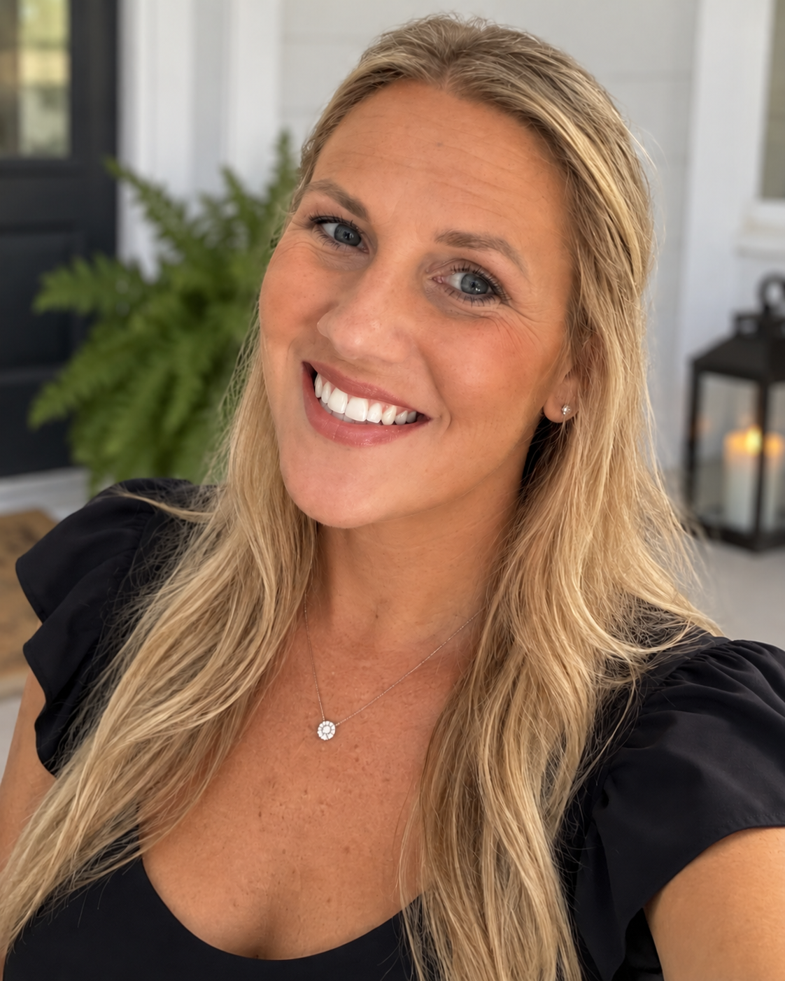 A woman with blonde hair and blue eyes smiling, wearing a black top, a necklace with a circular pendant, and pearl earrings. In the background, there is a potted plant, a lantern with a candle, and a door.