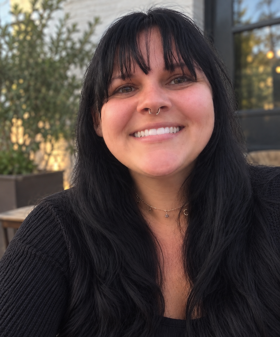 A woman with dark, long hair, light skin, and a septum piercing smiling outdoors during daytime, wearing a black top and layered necklaces, with greenery and a wooden structure in the background.