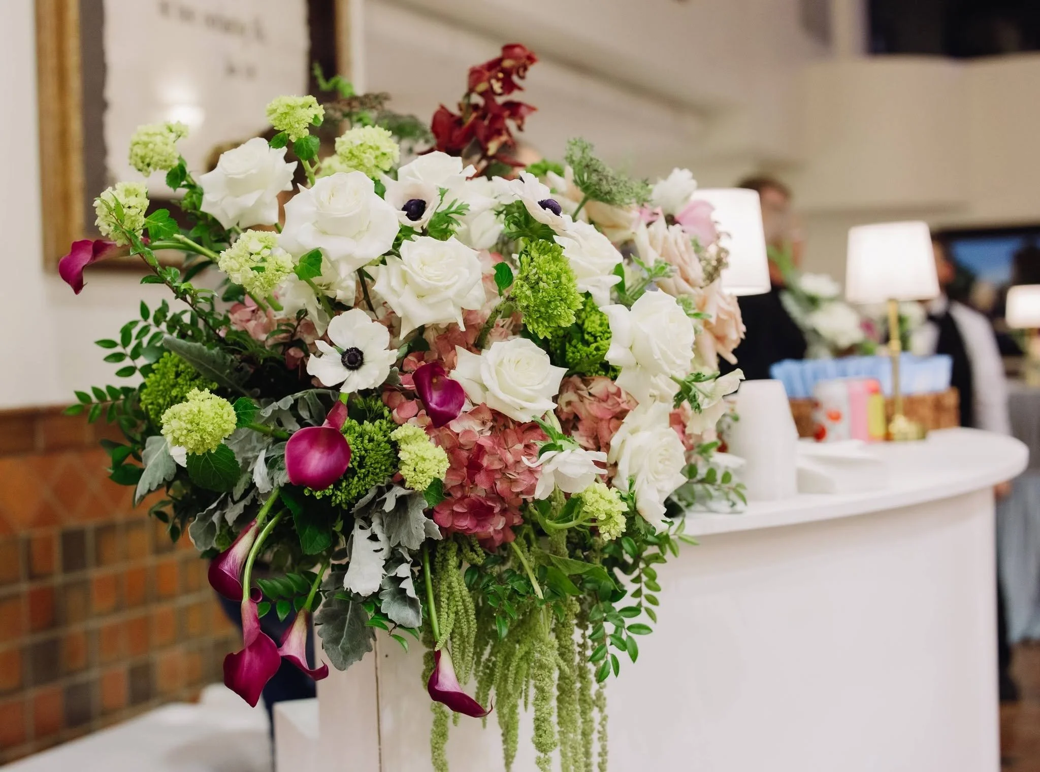 A large floral arrangement featuring white roses, pink calla lilies, green hydrangeas, and various green foliage, displayed indoors on a white table with lamps and blurred people in the background.