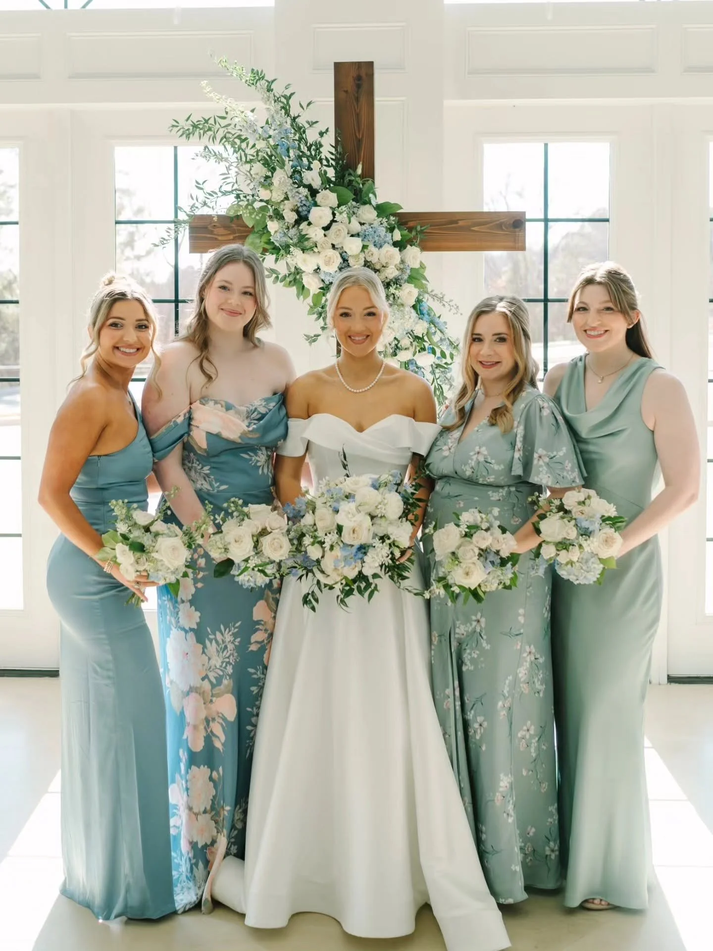 A bride and four women in matching blue floral dresses holding bouquets of white and blue flowers, standing in front of a large wooden cross decorated with white and blue flowers and green foliage, inside a bright room with large windows.