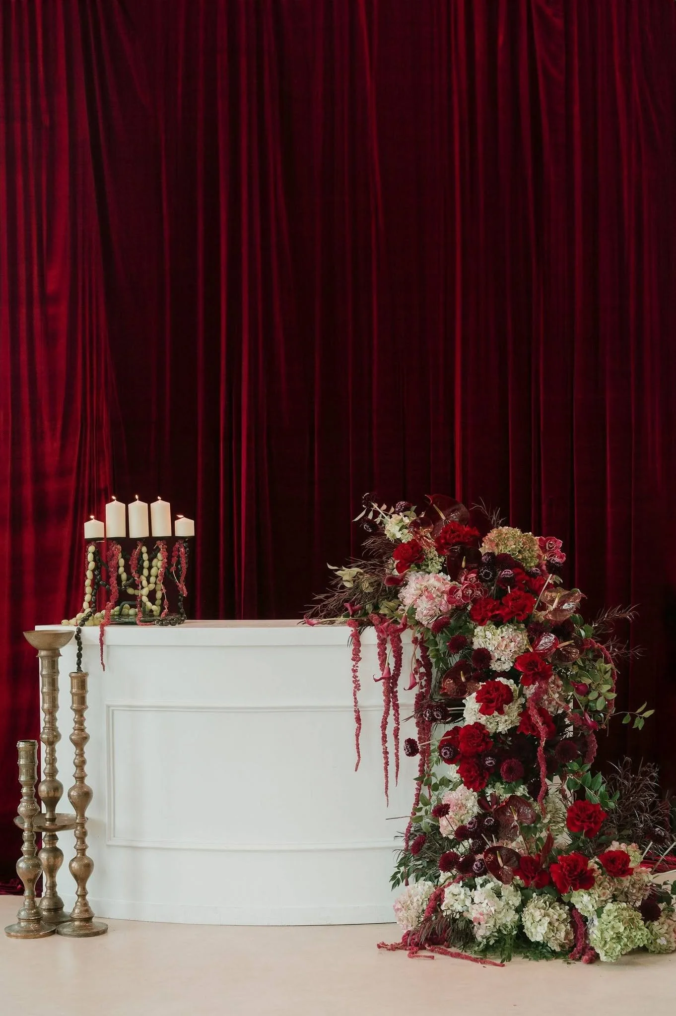 Elegant floral arrangement of red, white, and dark purple flowers cascading next to a white structure with candles and gold candle holders, set against dark red curtains.