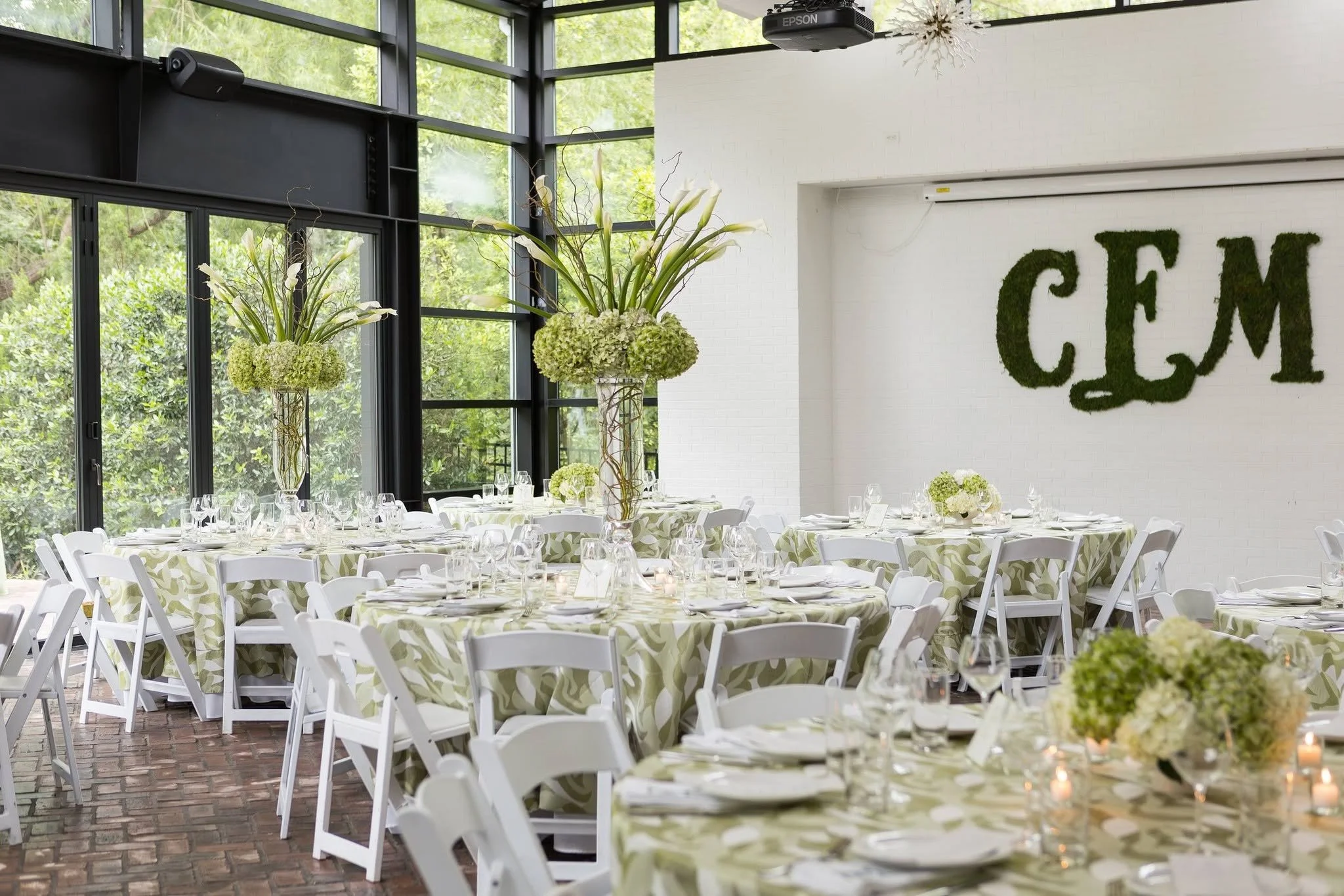 Decorated banquet hall with round tables covered in green leaf-patterned tablecloths, white chairs, floral centerpieces with white flowers, tall flower arrangements, large windows, and a white wall with a green moss-like sign.