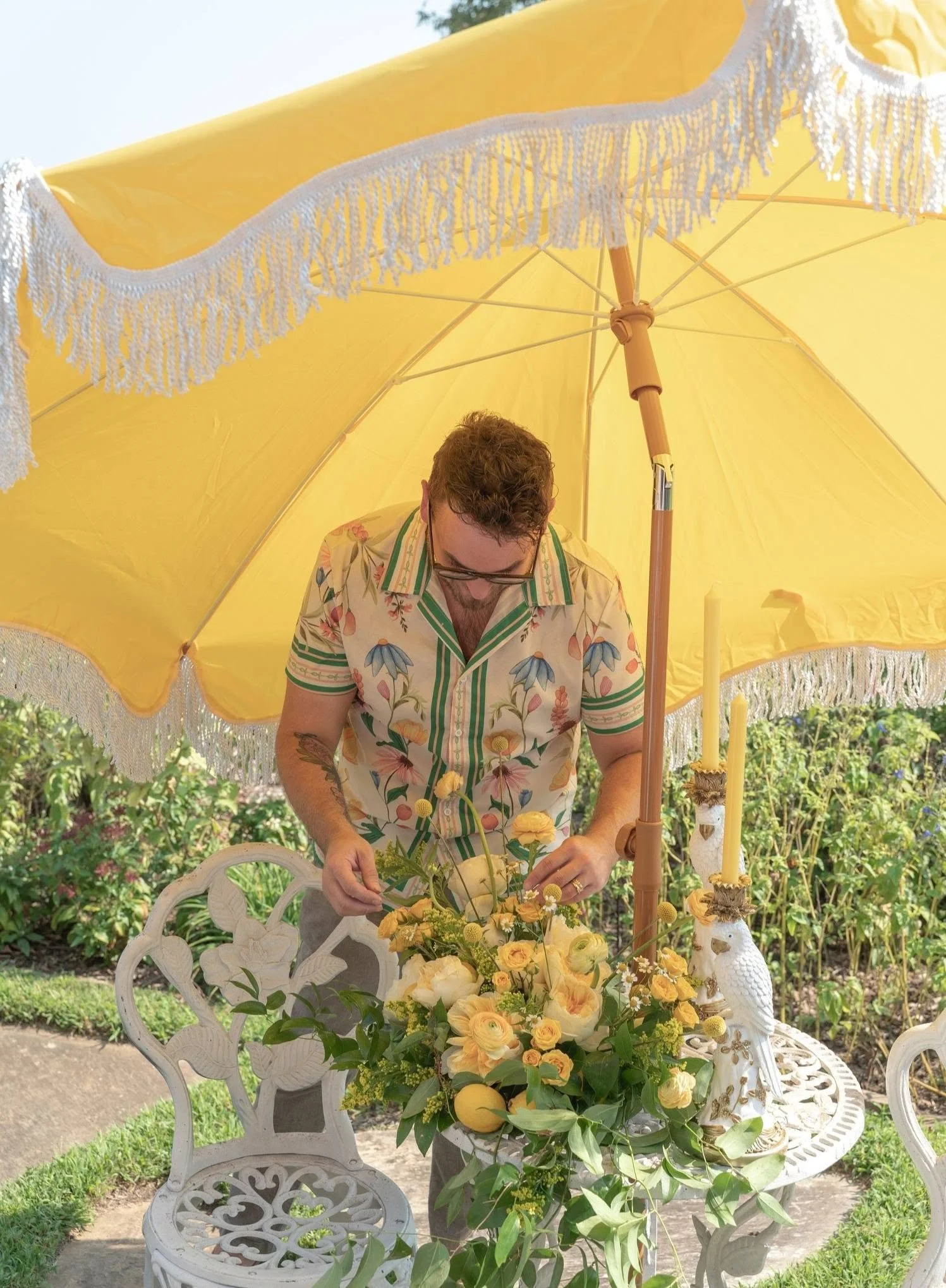 A man in vintage Hawaiian shirt arranging flowers on a table under a large yellow parasol with lace trim in a garden.